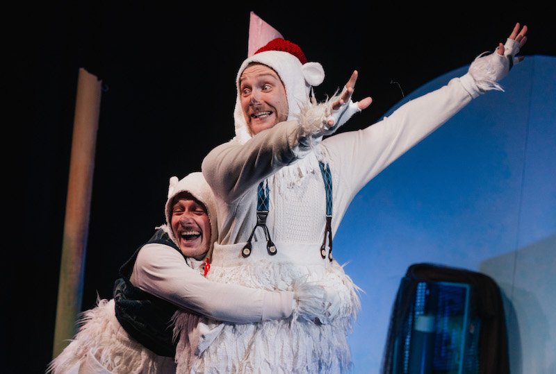 Two smiling performers in fluffy white bear costumes pose playfully on a dark blue stage. One swings his arms and the other hugs him.