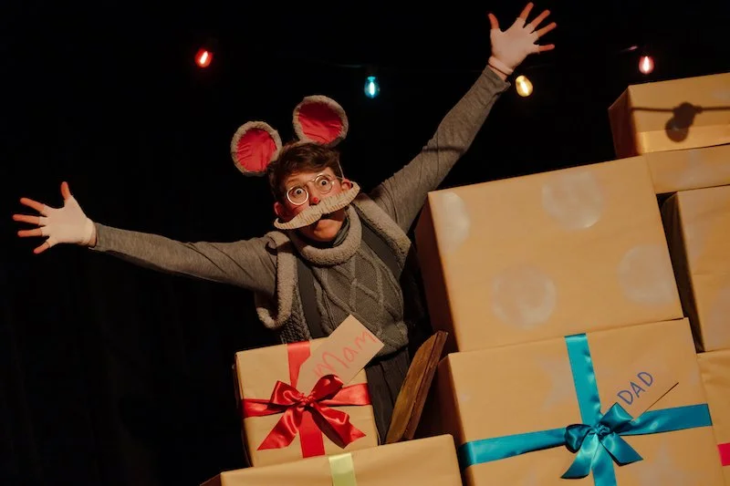 A performer dressed as a grey mouse with big pink ears and a large moustache leans over wrapped presents with arms wide.