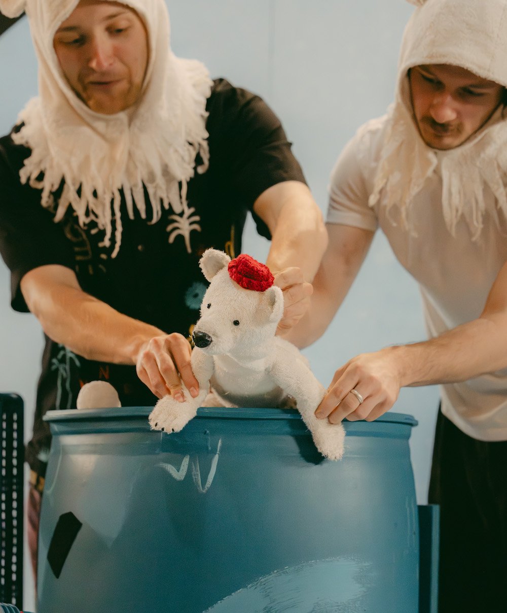 Two figures (one wearing a black t-shirt and other a white t-shirt) lift a fluffy white bear out of a blue plastic barrel. the bear wears a red hat and appears to be looking at it's surroundings