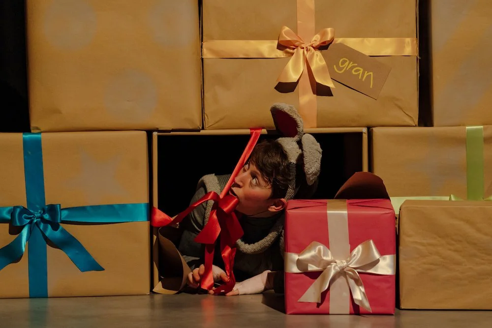 A performer wearing grey mouse ears pops out of a hole in a stack of brown paper presents tied with colourful ribbons.