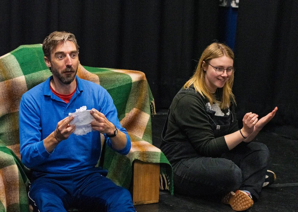 A person wearing a blue sweatshirt sits on a chair covered in a checkered green and brown blanket and holds a crumpled piece of paper in front of them. A person with blonde hair wears black and sits in the floor next to them