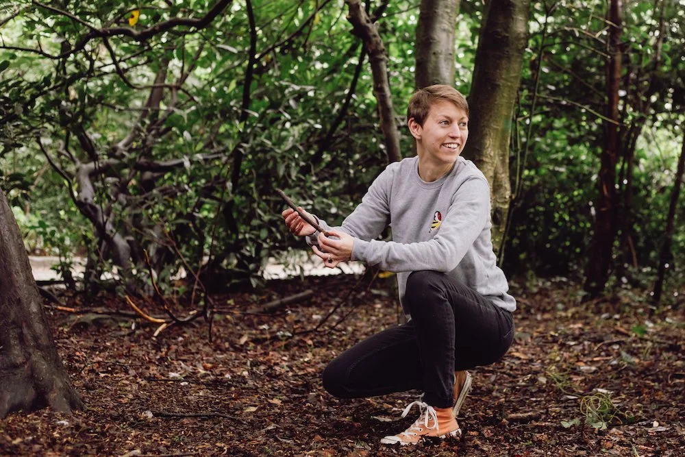 A short-haired person in a grey sweatshirt crouches in woodland, smiling with arms outstretched while holding a small stick.