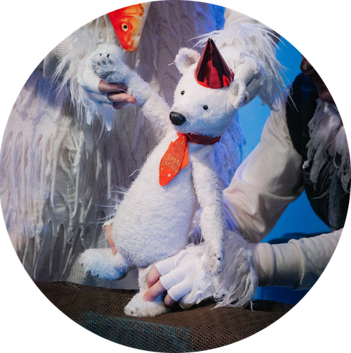 A white stuffed bear in a red hat is moved across a surface by two performers in fluffy white costumes guiding its limbs.