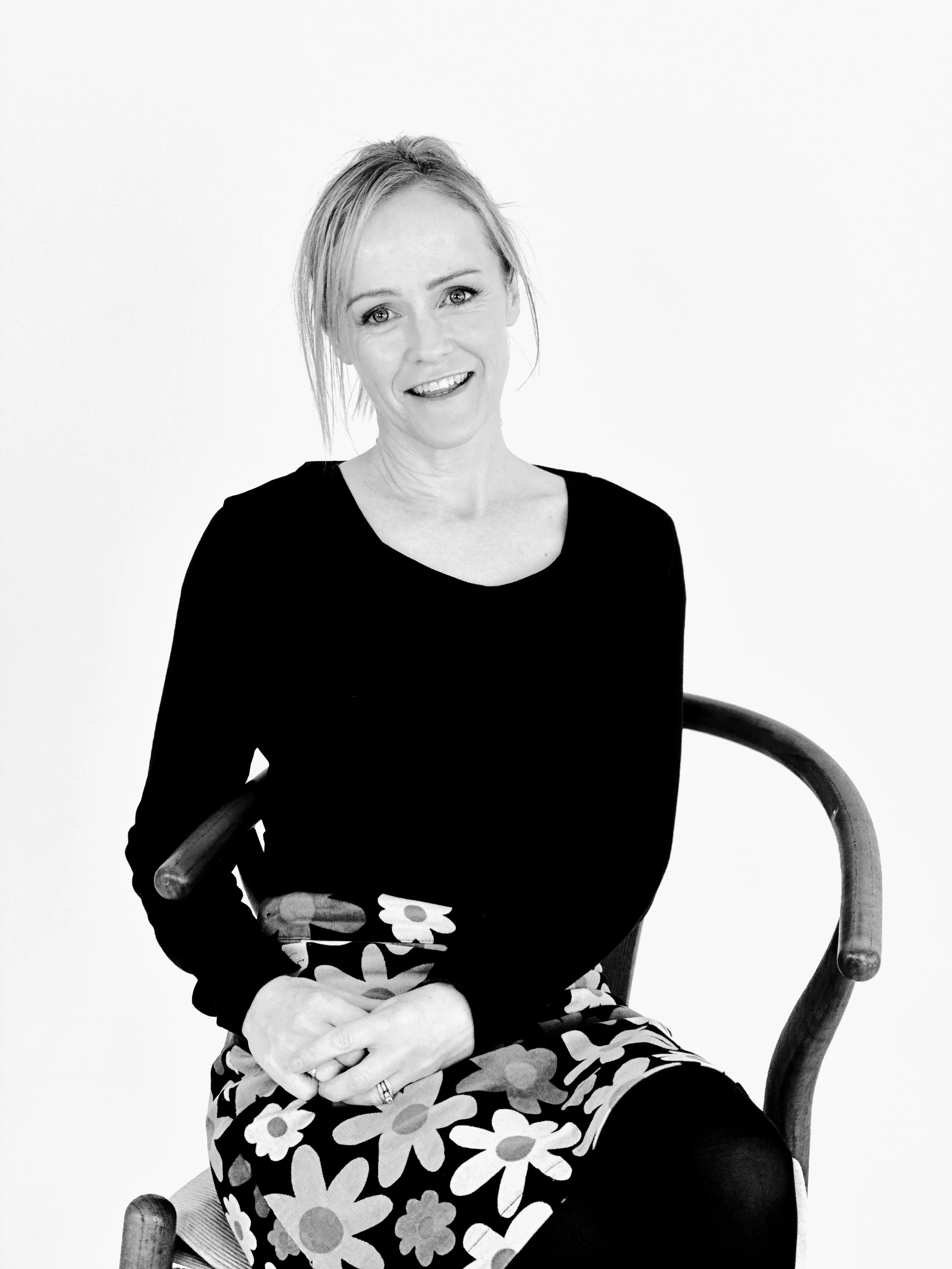 A woman sitting on a wooden chair, smiling at the camera, in a black long-sleeve top and a floral skirt, in a studio setting.