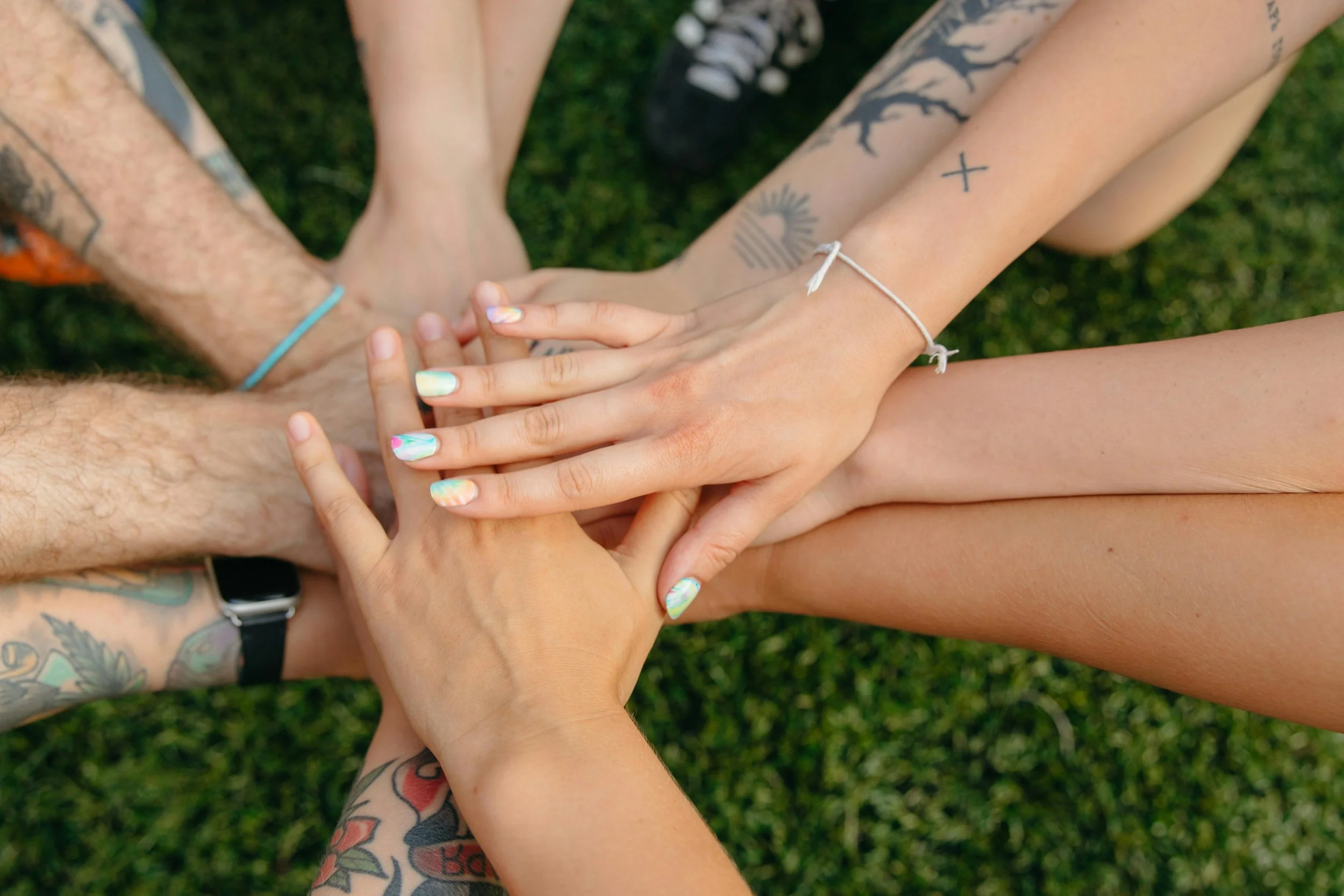 Multiple arms and hands of diverse individuals stacked together in a circle, showing tattoos and different skin tones, with a grassy background.