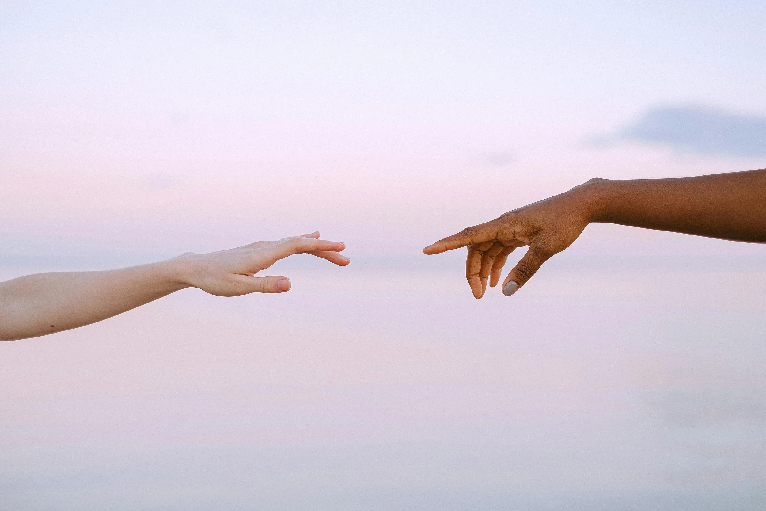 Close-up of two hands reaching toward each other against a pastel sky.