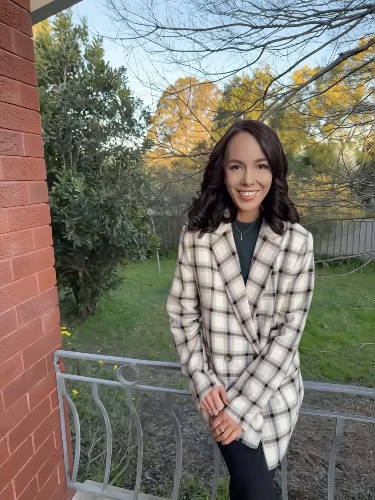 A woman with dark, wavy hair and a bright smile standing outdoors on a patio or porch, wearing a checkered blazer, with trees and a fenced yard in the background during fall.
