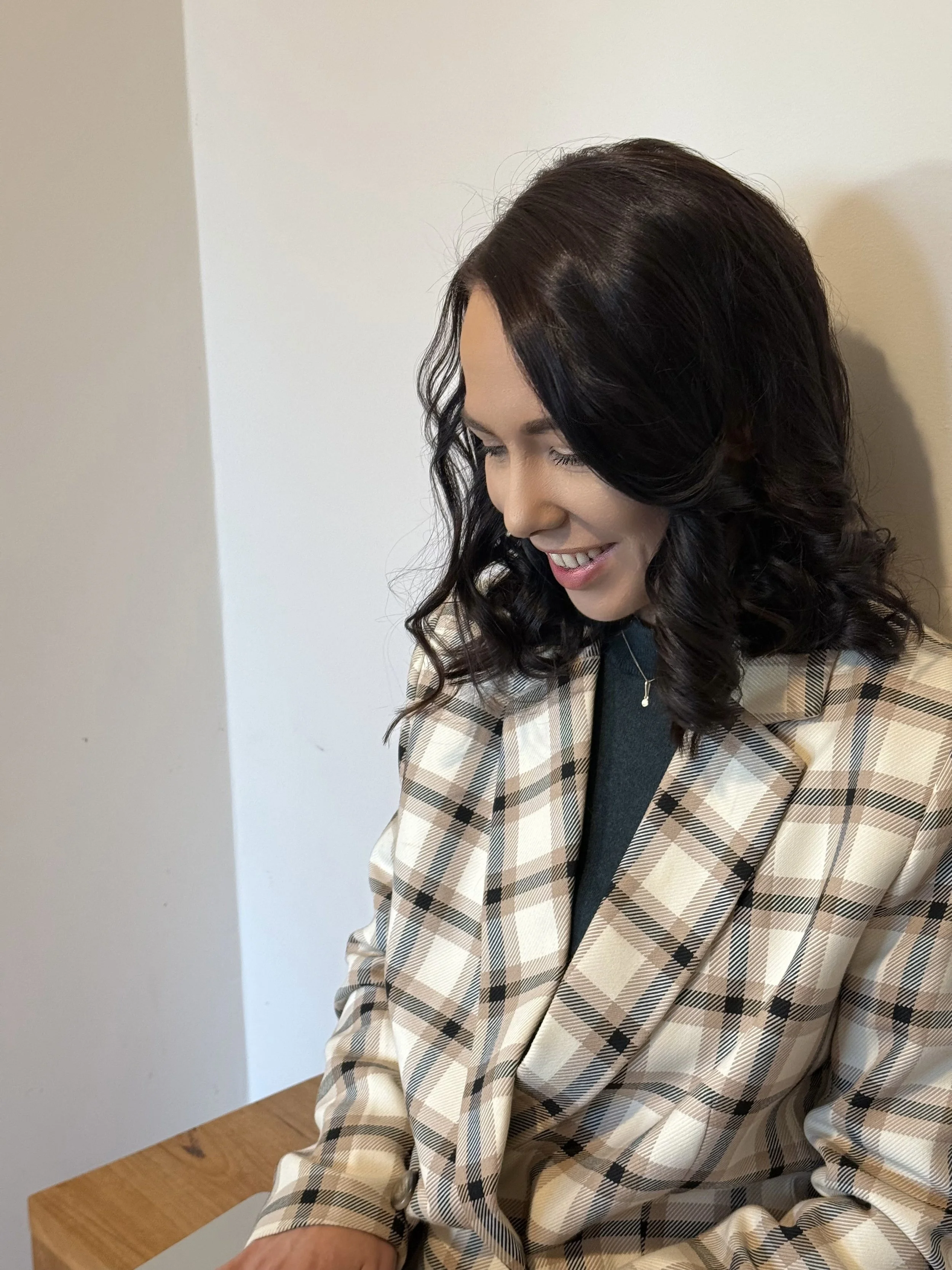 A woman with dark, wavy hair wearing a beige plaid blazer and black top, smiling and looking down, seated next to a white wall.