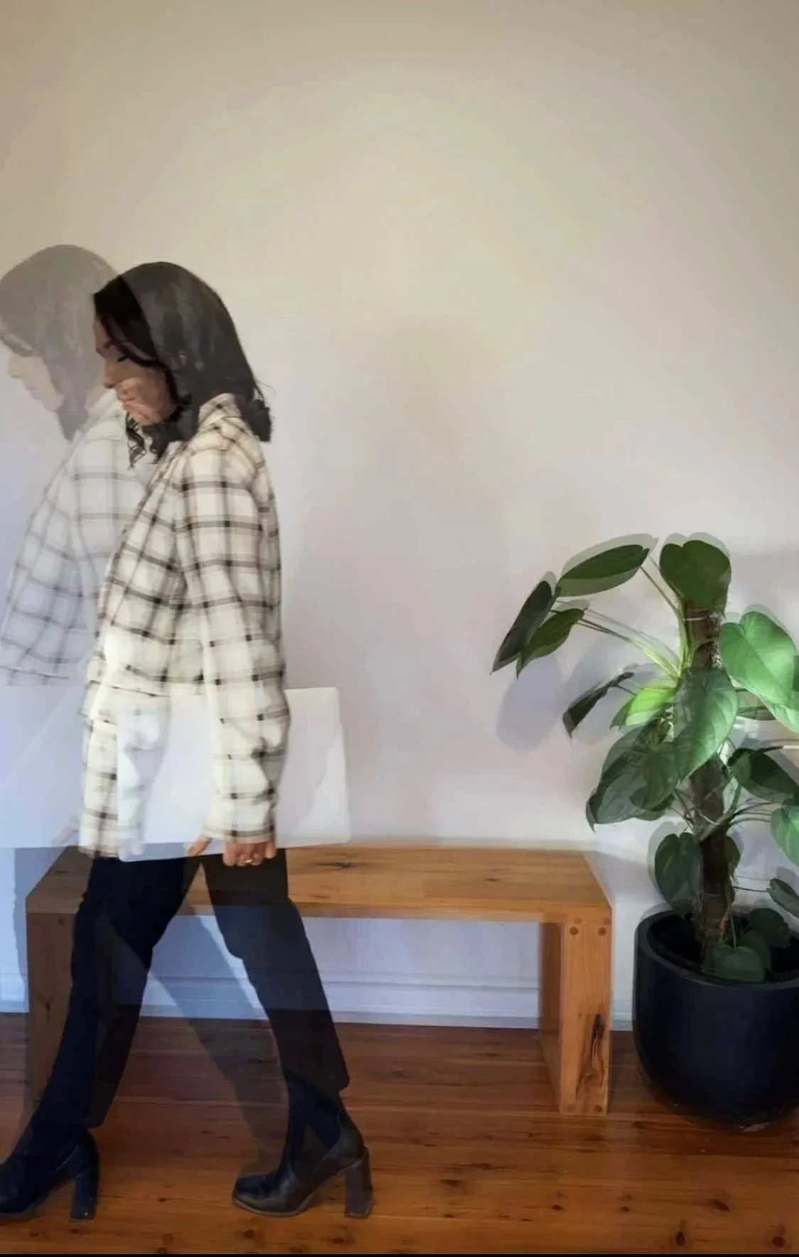 A woman in a plaid coat carrying a white bag, walking past a wooden bench and a potted plant against a plain wall.