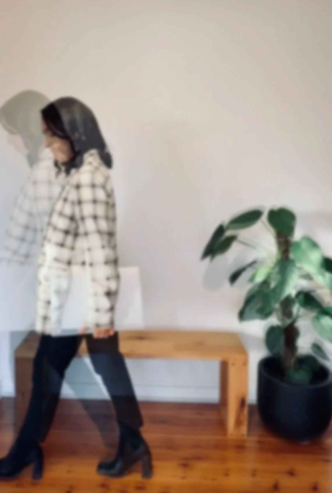 A woman dressed in a checkered outfit with high-heeled boots walking past a large potted plant in a room with wooden flooring and a simple wooden bench against a plain white wall.