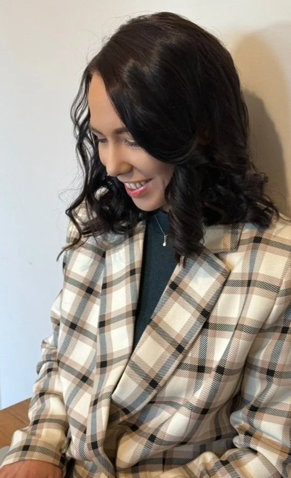 A woman with dark, wavy hair smiling and looking down, wearing a beige plaid blazer over a dark top, against a plain background.