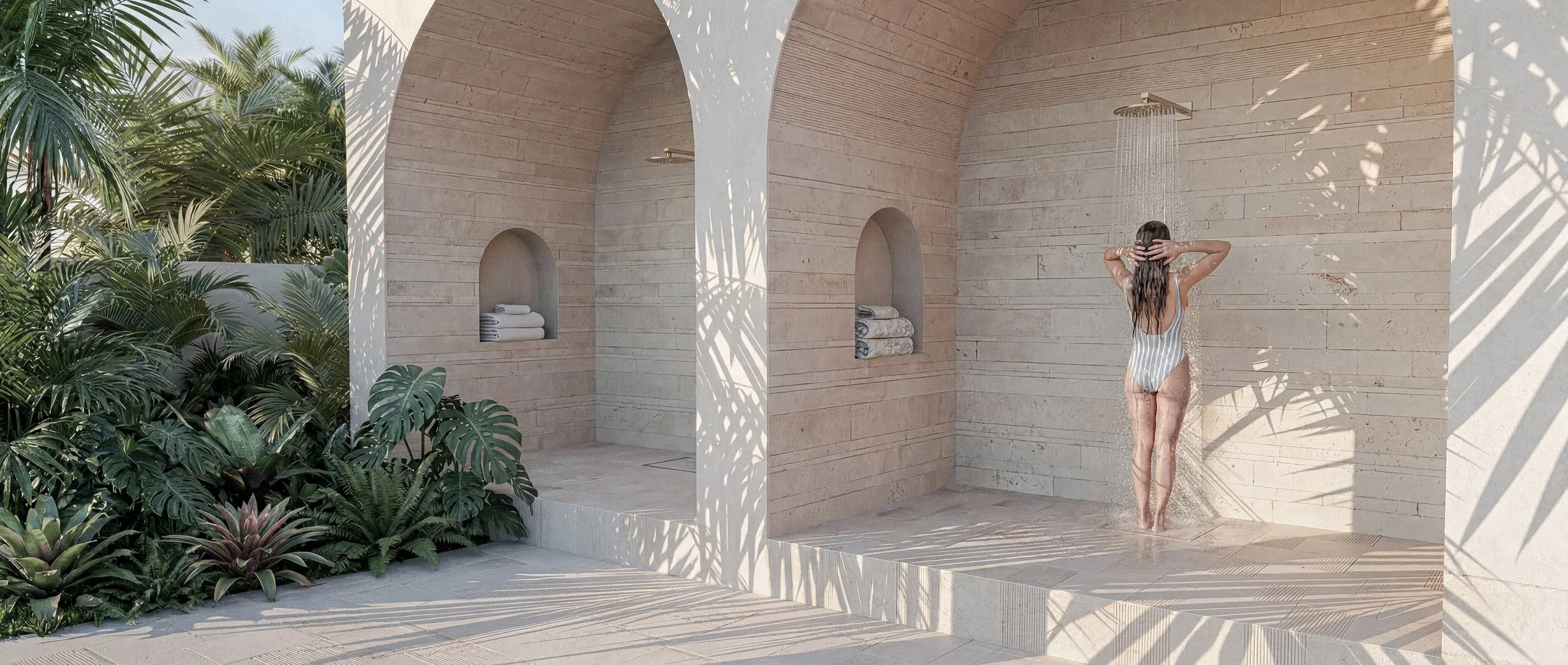 An outdoor shower area with a stone tile floor, an orange plaster wall with metal shower heads, and tall palm trees casting shadows on the ground.
