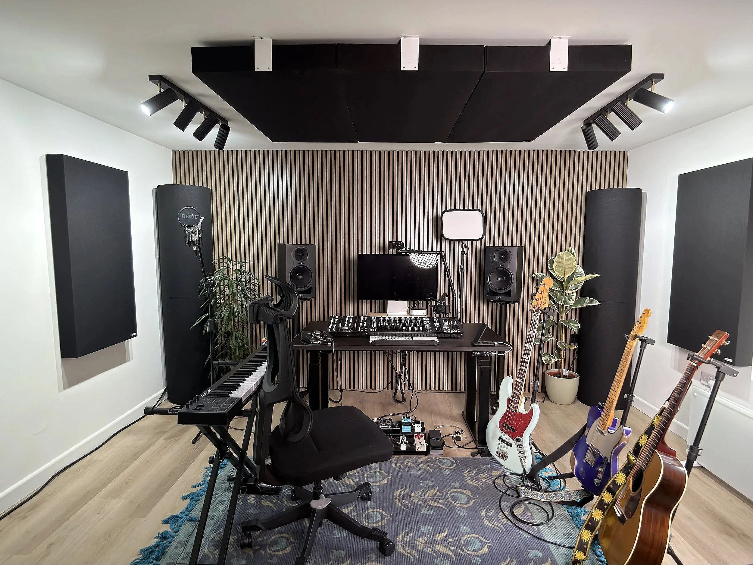 Musician's recording studio with guitars, keyboard, mixing console, studio monitors, microphone, TV, and acoustic panels.