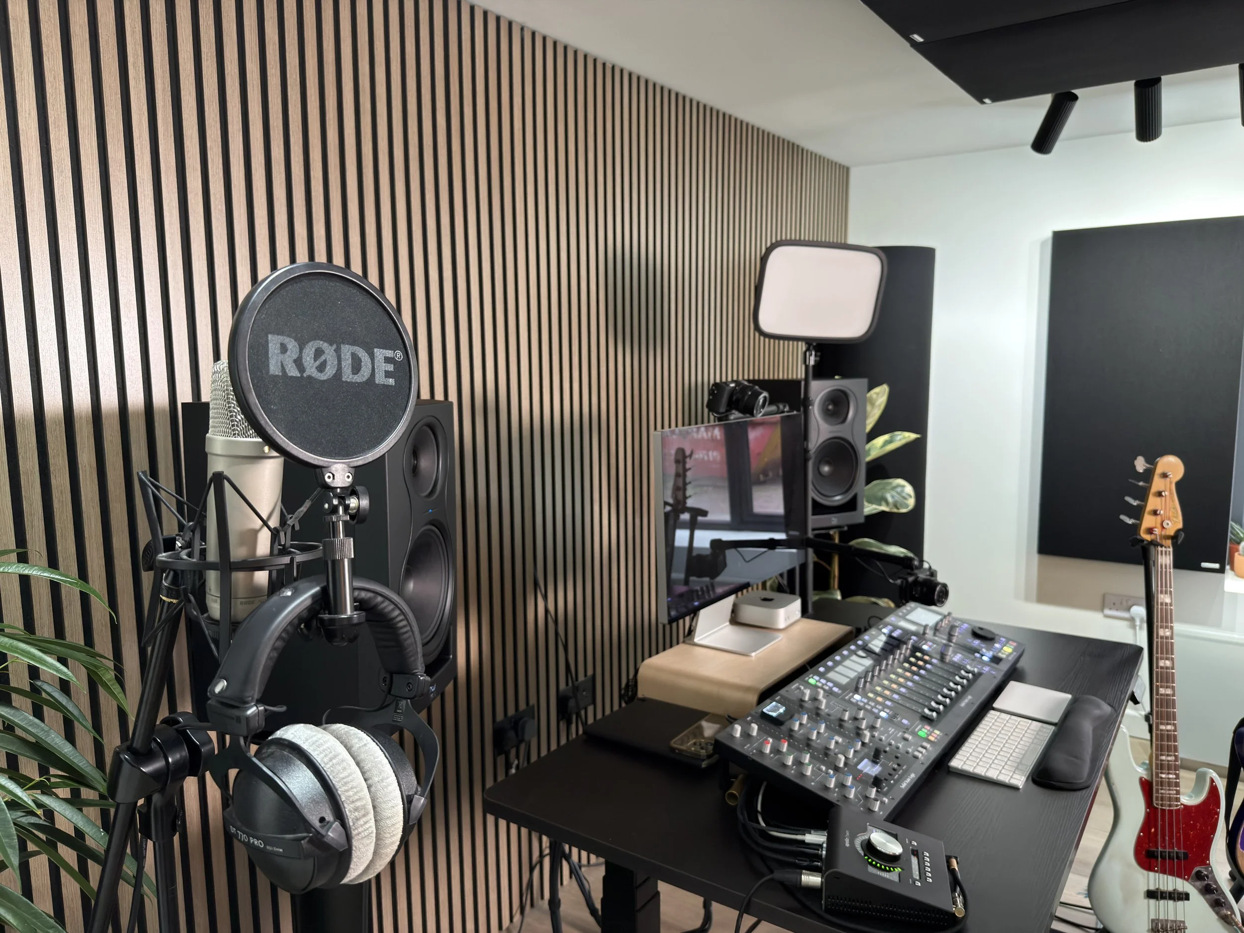 A music recording studio featuring a microphone with a RØDE pop filter, headphones hanging on a stand, studio monitors, a digital audio mixer, a computer monitor, a keyboard, a guitar, and acoustic treatment panels on the wall.