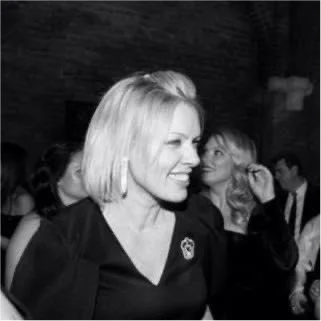 Black and white photo of a smiling woman with shoulder-length blonde hair at a social event, with other people in the background.
