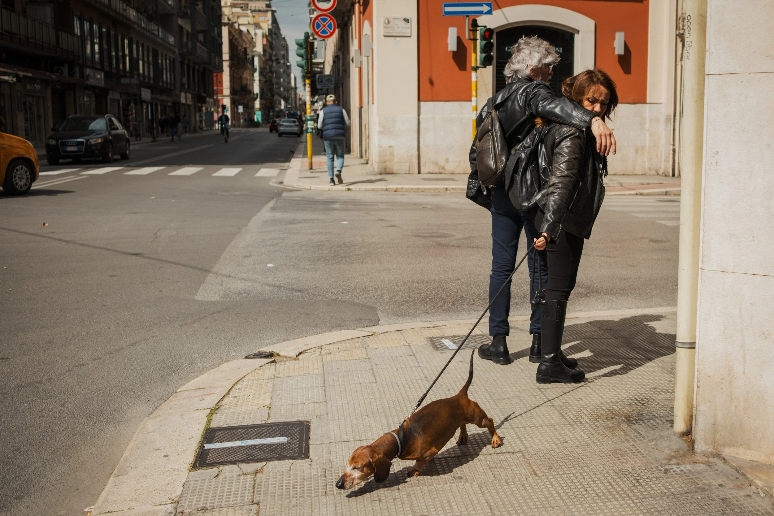 Two women, one with gray hair and the other with reddish hair, standing on a city sidewalk. The woman with gray hair has her arm around the other woman. They are wearing leather jackets. The reddish-haired woman is walking a brown dog on a leash, sni