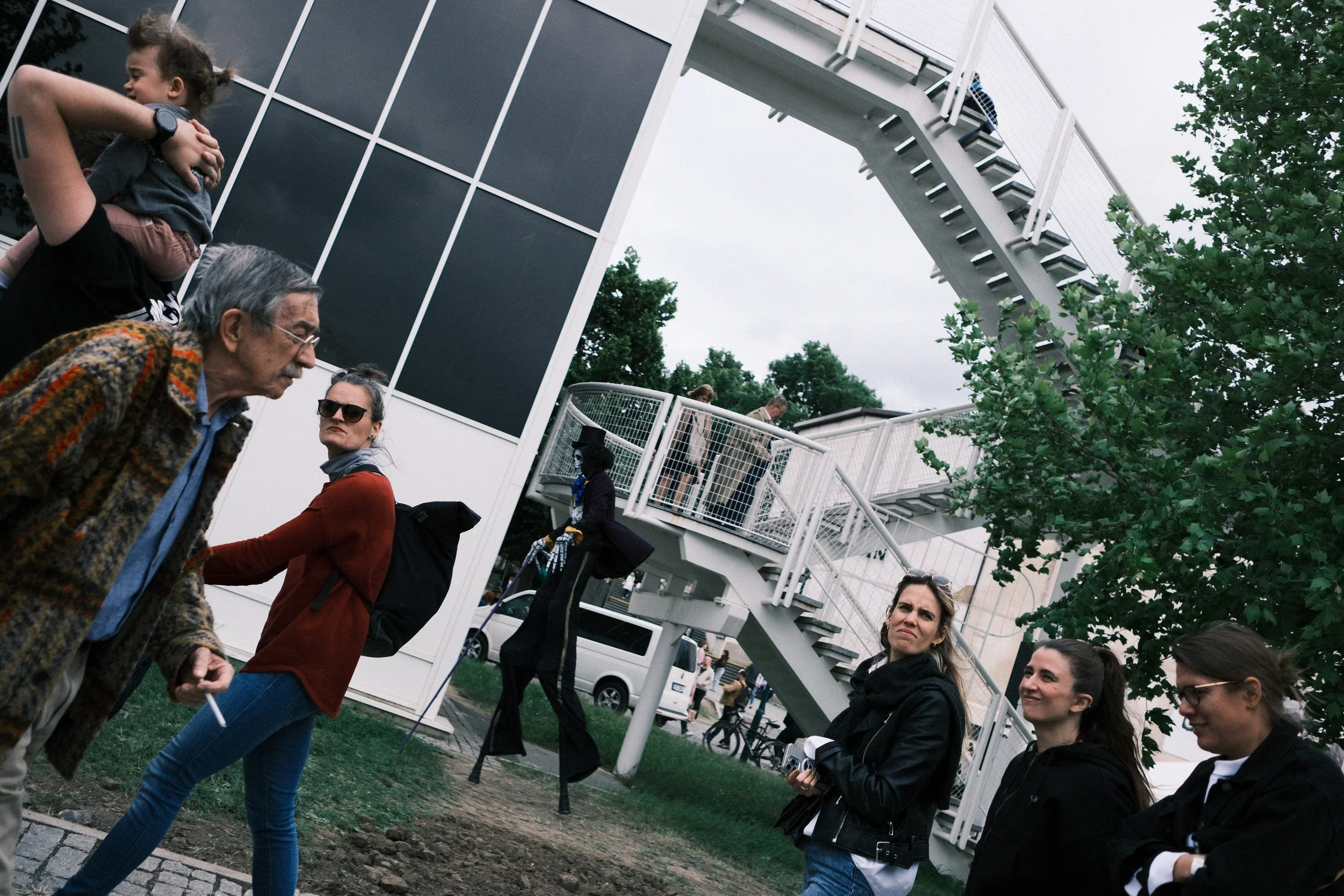 A group of people standing near an outdoor staircase, with some engaged in conversation and others looking around. The scene is set outside a modern building with large windows, and there is a tree and some cars in the background. It appears to be a 