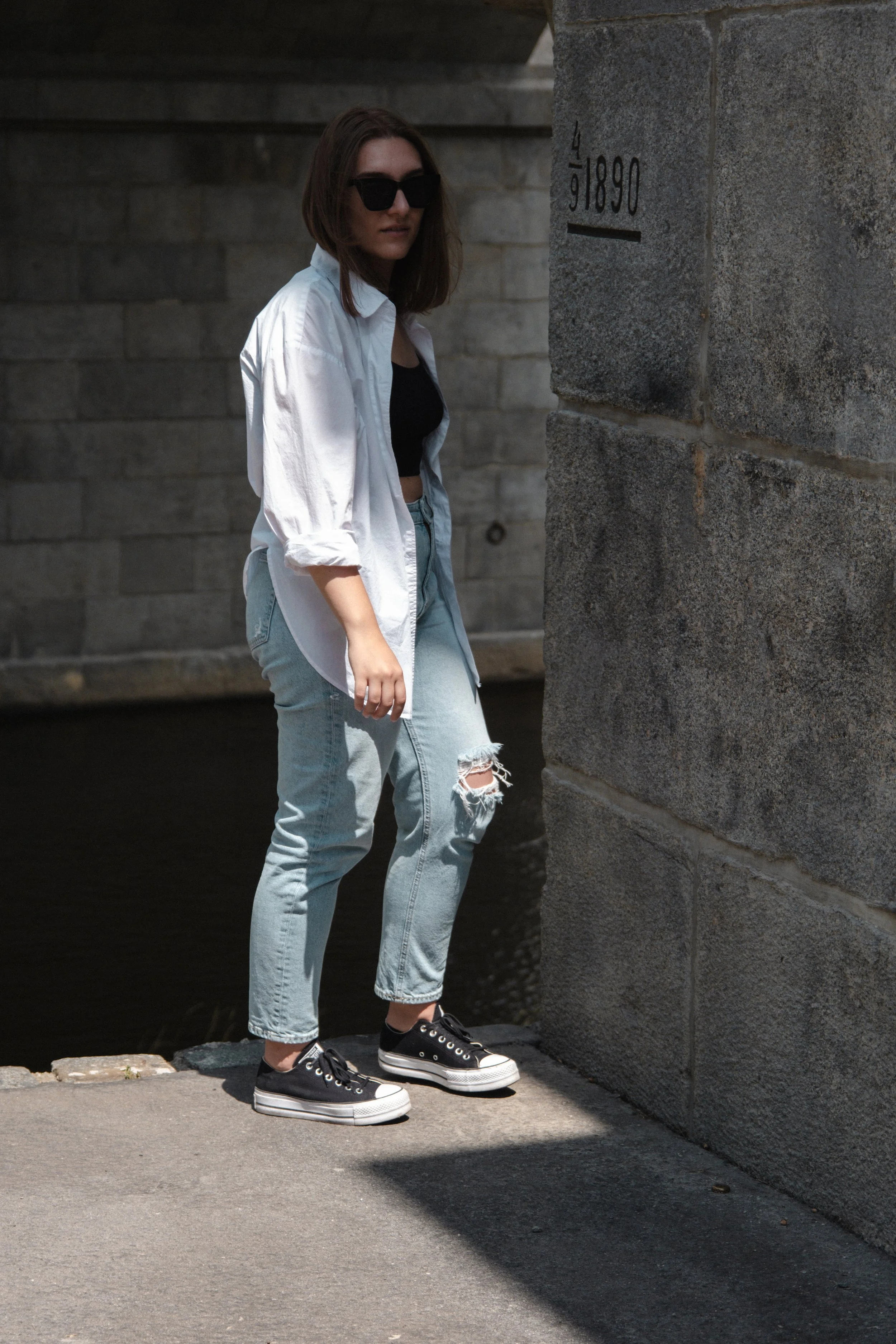 A young woman with shoulder-length brown hair wearing sunglasses, a white button-up shirt over a black top, ripped light-wash jeans, and black sneakers standing near a stone wall by a waterway.