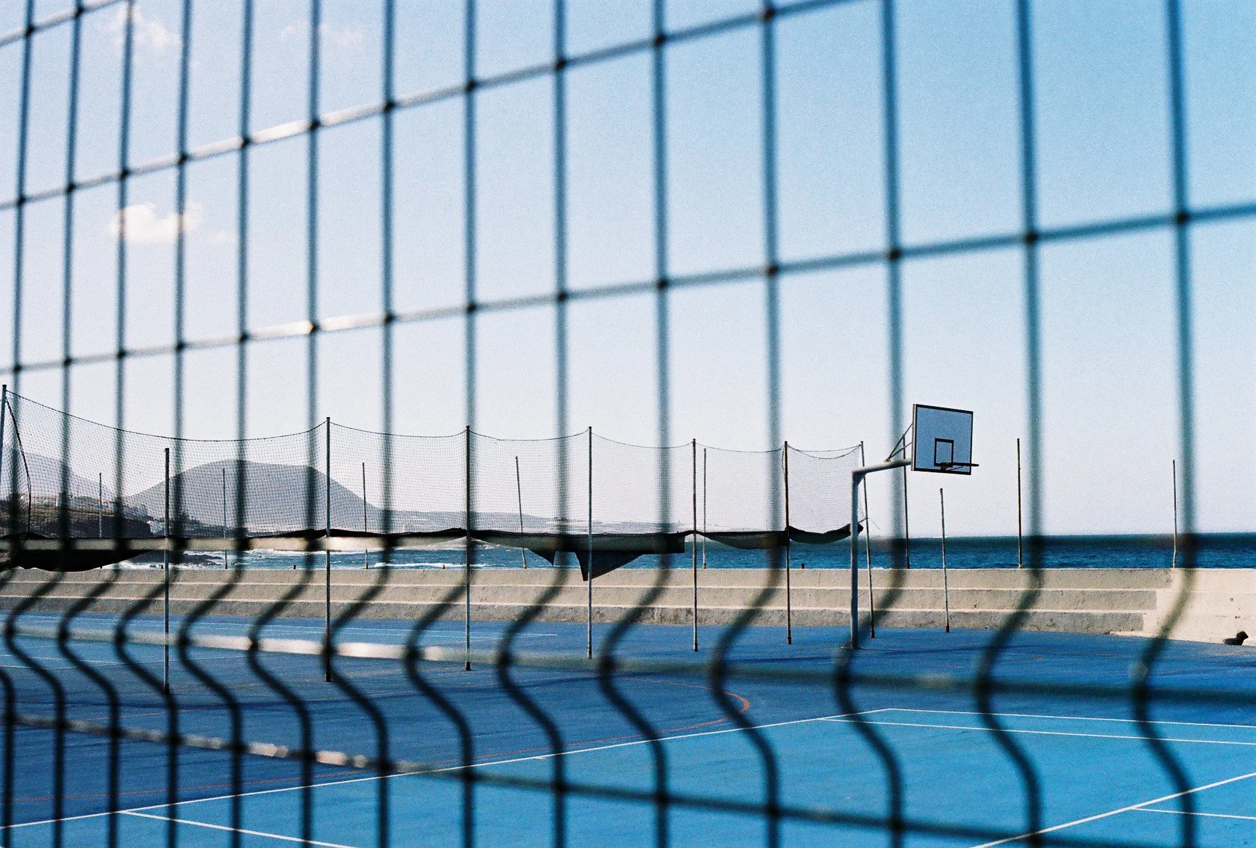 An empty basketball court with a hoop, viewed through a chain-link fence, near the ocean with mountains in the background.