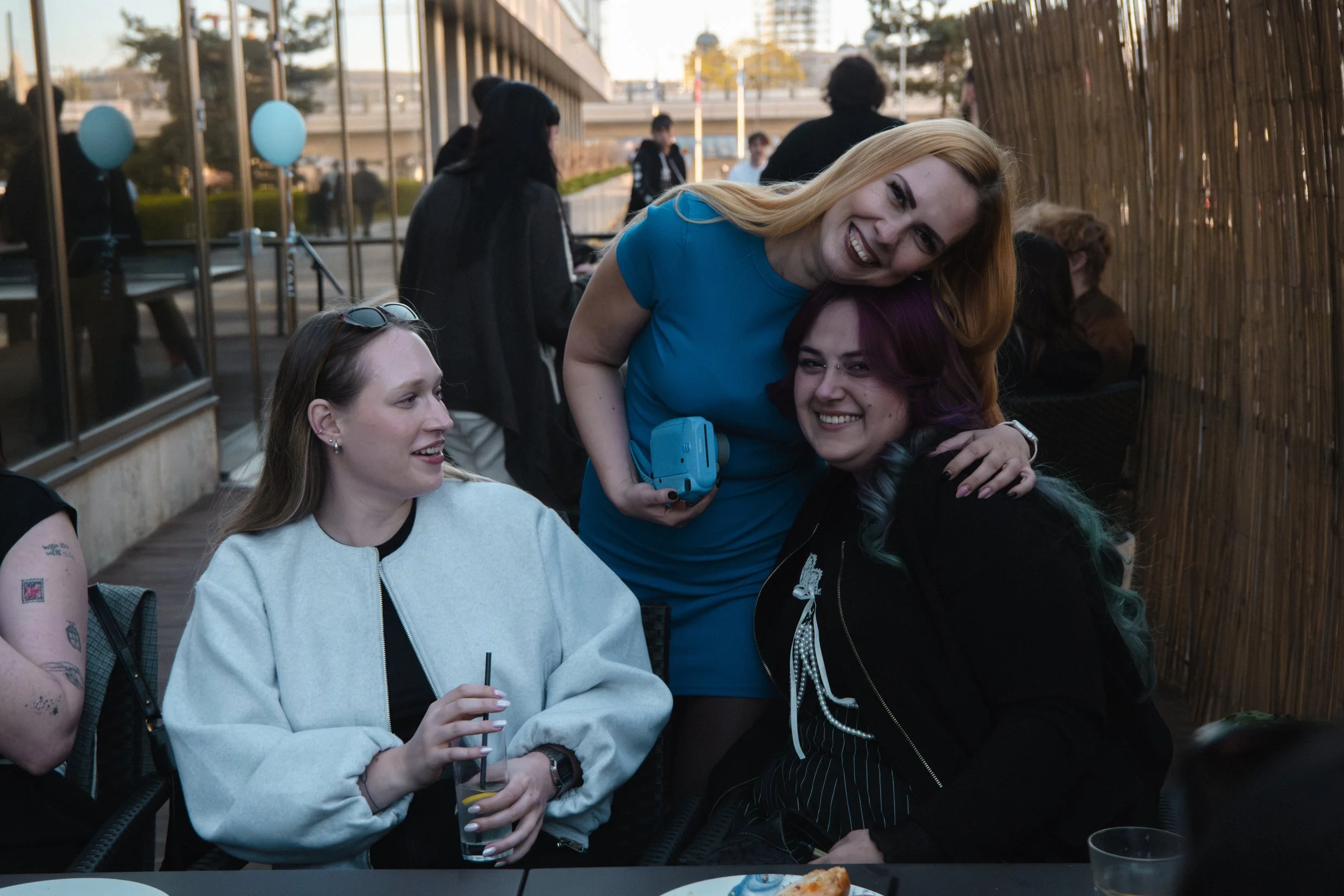 Three women sitting and standing at a table at an outdoor gathering, smiling and interacting. One woman with blonde hair and a blue dress stands behind two women seated, hugging one and holding a blue object. The woman on the left has long brown hair