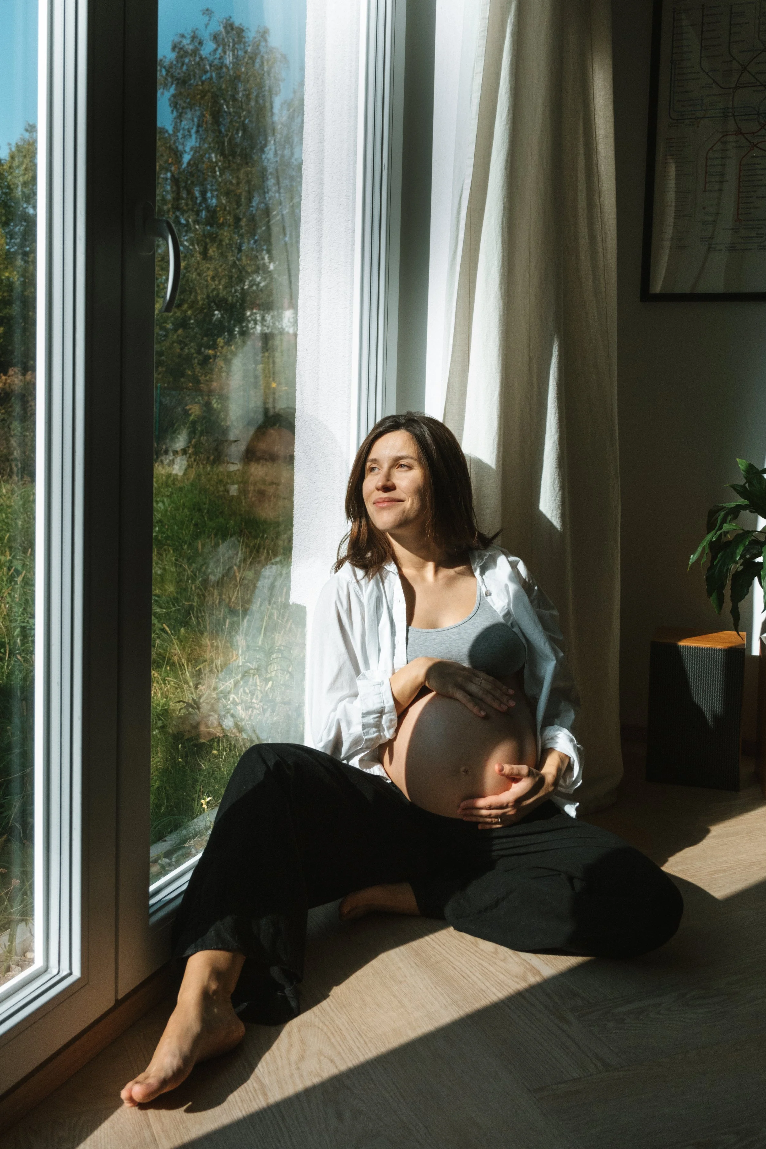Pregnant woman sitting on the floor by a glass door, basking in sunlight with a serene expression.