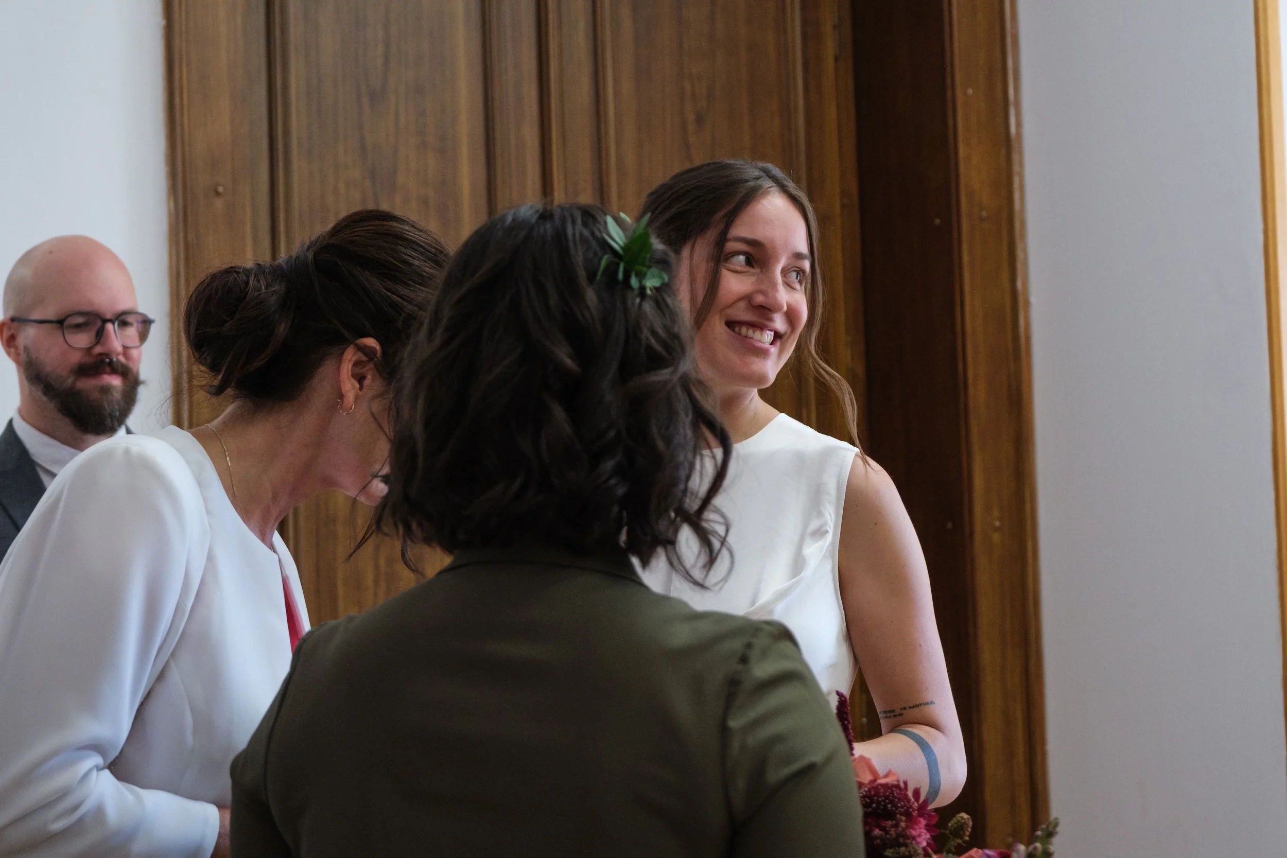 A group of people at a formal event, including a smiling woman with long brown hair in a white dress holding a bouquet of flowers, standing by a wooden wall.