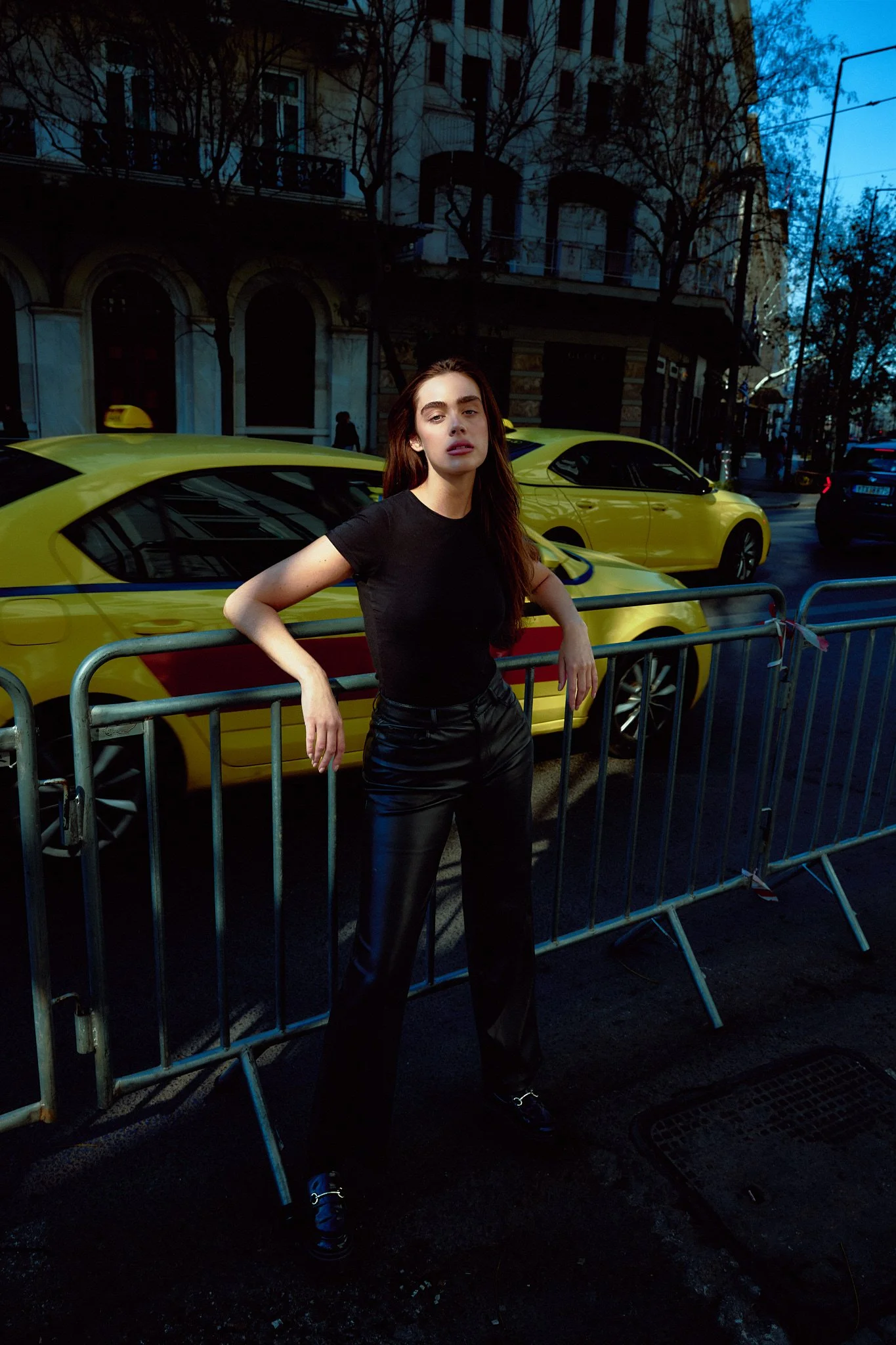 A young woman with long brown hair wearing a black shirt and black leather pants standing behind a metal barricade on a city street at dusk, with yellow taxis and buildings in the background.