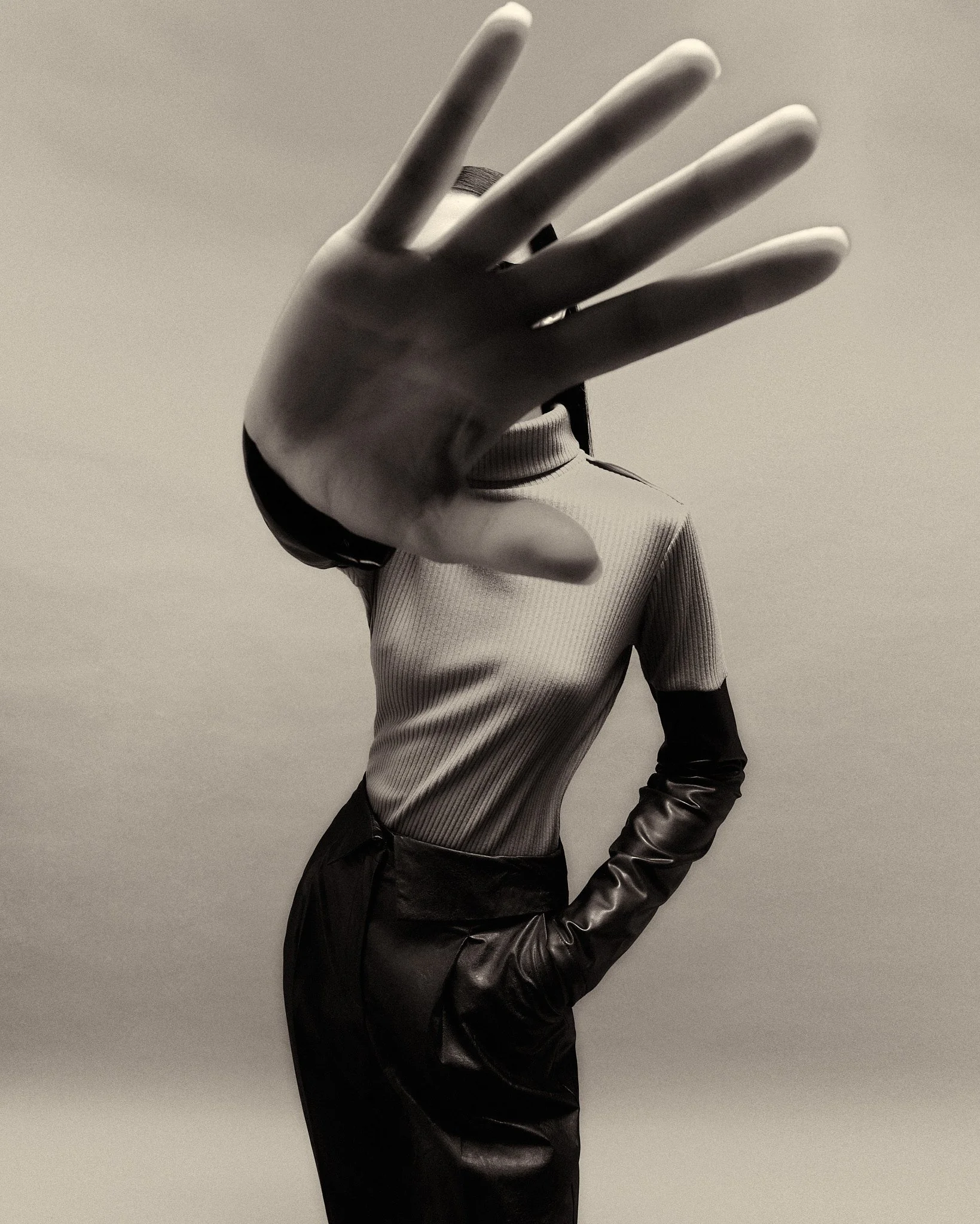A person in a turtleneck top and leather pants extending their hand toward the camera, obscuring their face.
