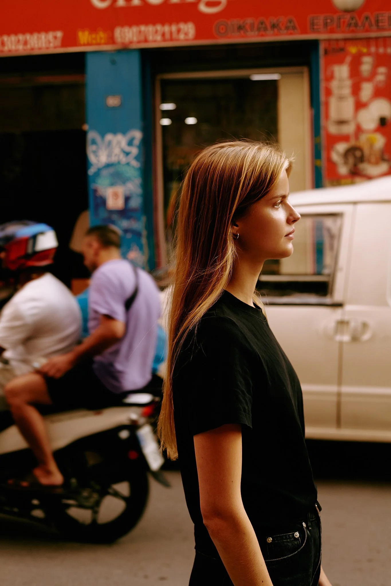 A young woman with long, light brown hair standing on a street at night, wearing a black t-shirt, with a colorful storefront and a white vehicle in the background.