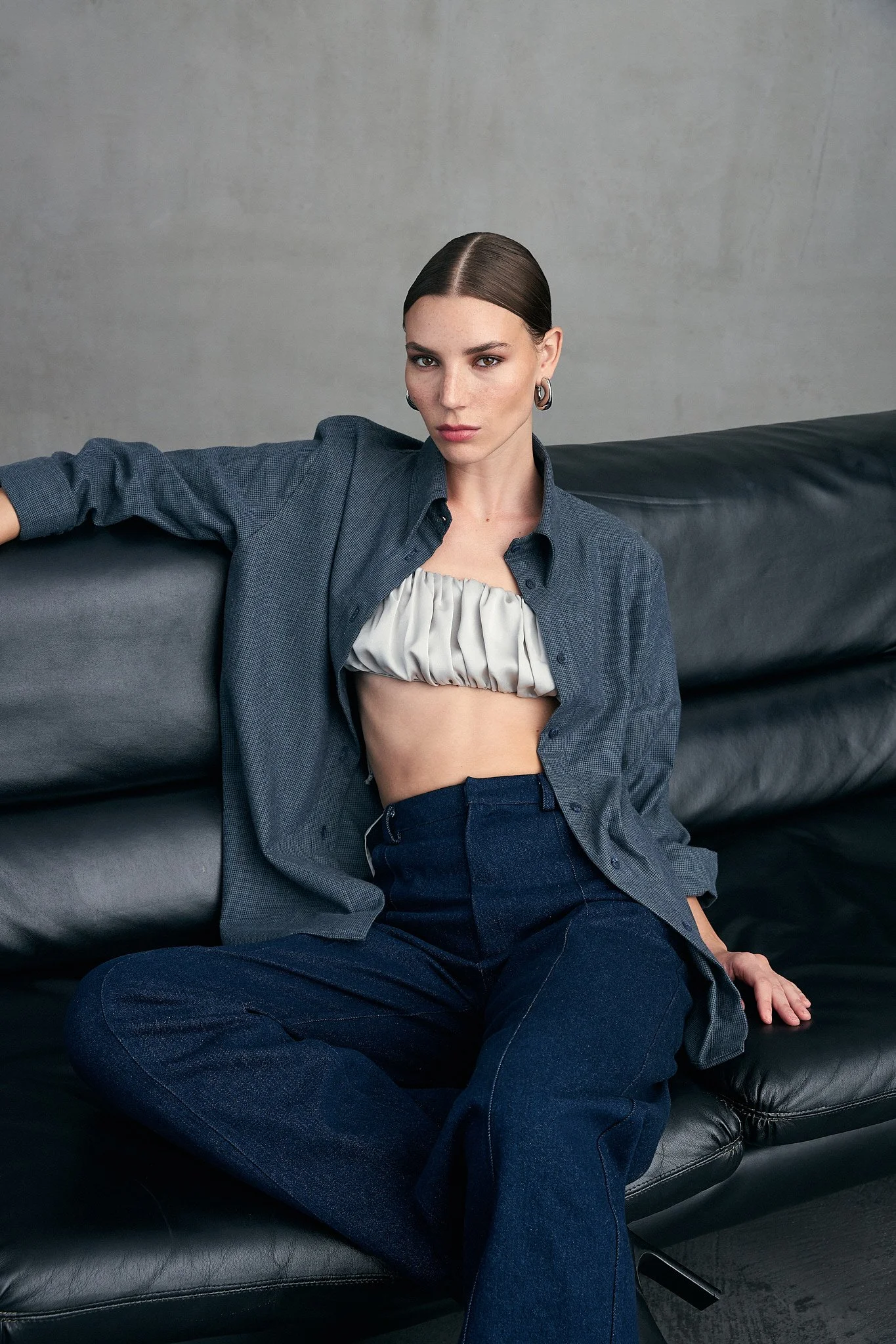 A woman with straight brown hair sitting on a black leather couch against a plain gray wall, wearing a gray button-up shirt, revealing a ruffled gray crop top, and dark blue jeans.