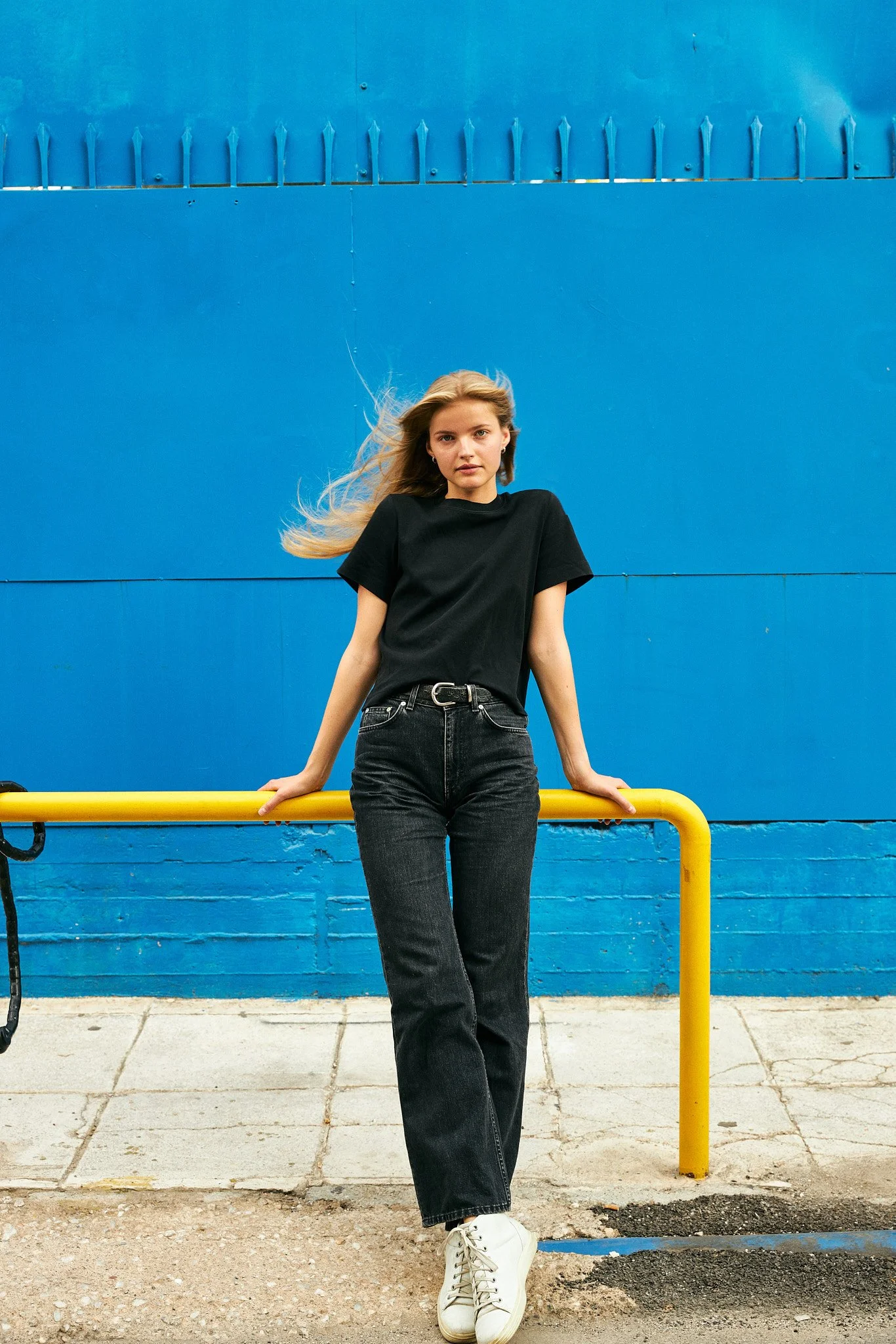 Young woman with long blonde hair wearing a black t-shirt, black jeans, white sneakers, standing against a blue wall with her hands on a yellow railing.