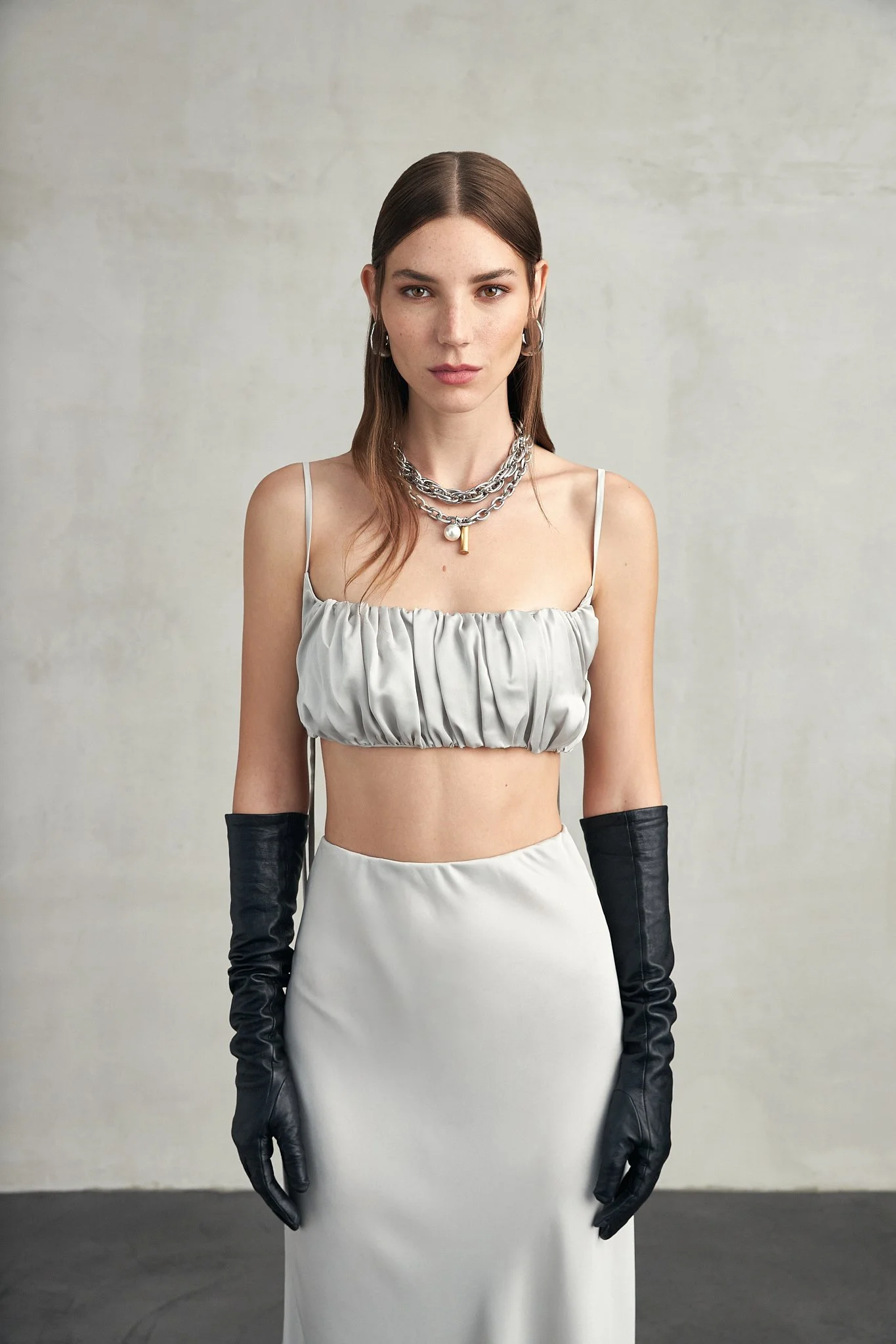 A young woman with light skin, brown hair, and hoop earrings is standing against a plain wall. She is wearing a silver top, a long white skirt, black gloves, and layered silver necklaces.
