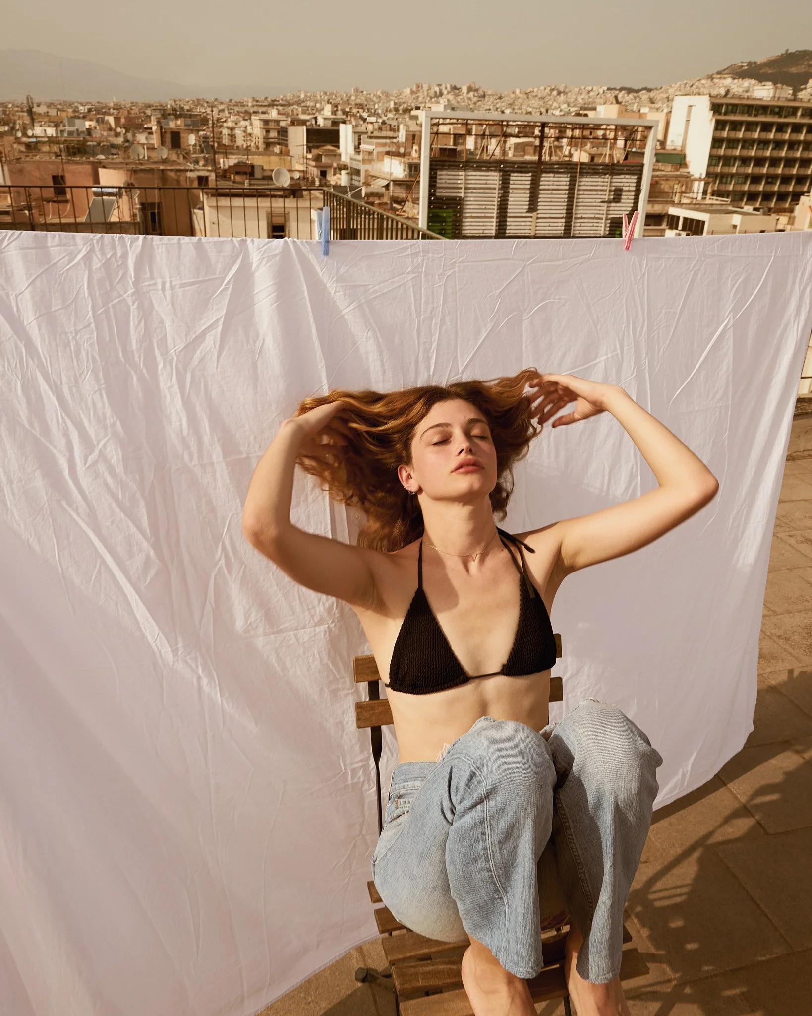 A woman with red hair sitting on a wooden chair on a rooftop, with a plain white sheet behind her, cityscape and mountains in the background, flipping her hair back with her eyes closed, wearing a black bikini top and light blue jeans.