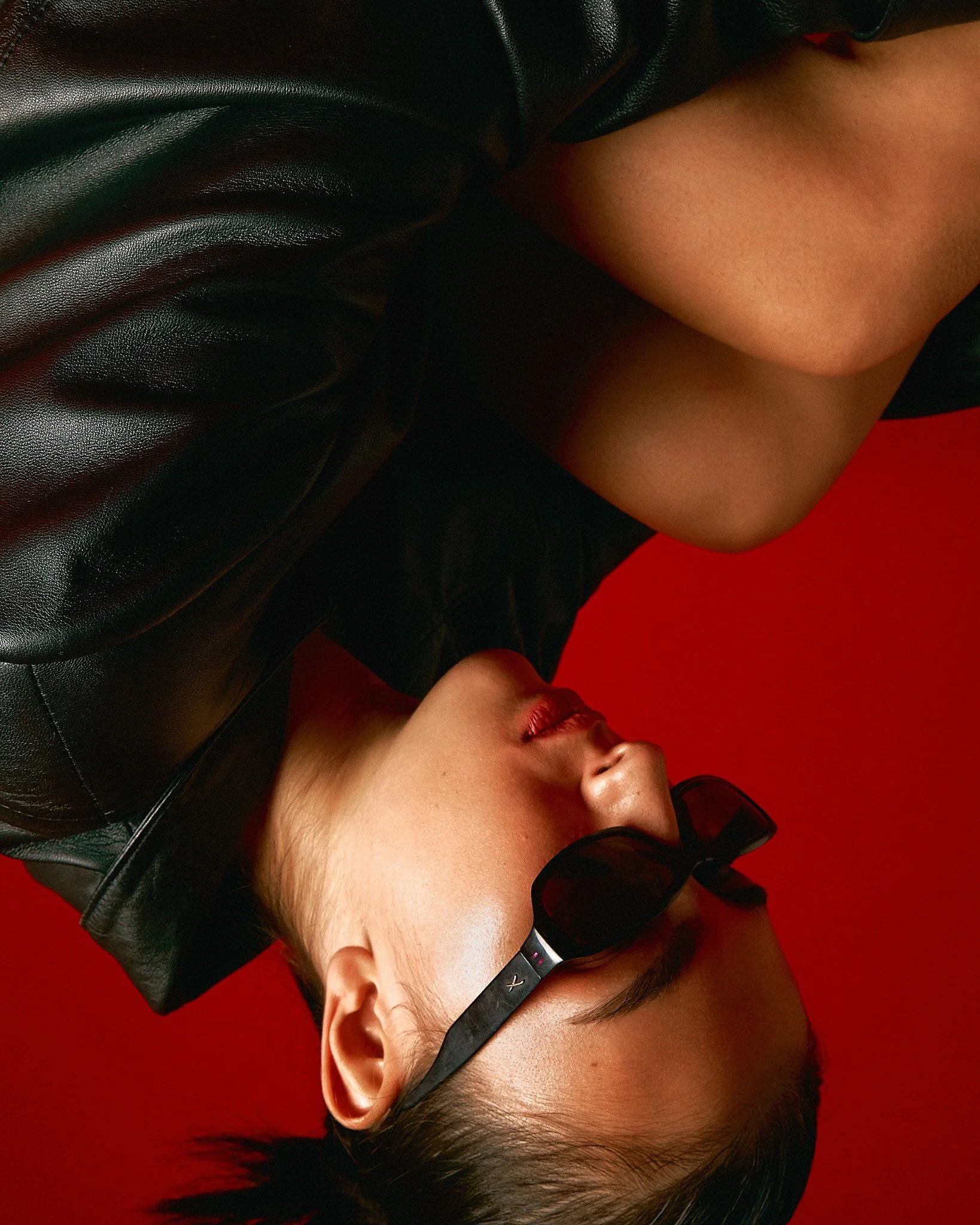 A young woman with dark hair, sunglasses, and a black leather jacket, lying on a red background with her head tilted back.