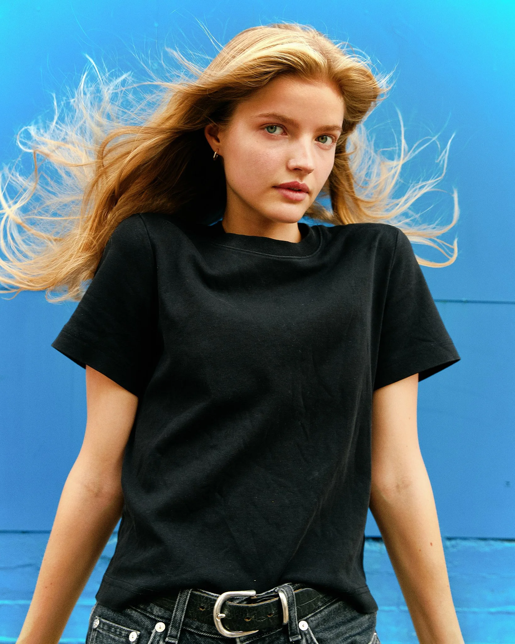 A young woman with long, blonde hair flowing in the wind, wearing a black t-shirt and black jeans with a belt, standing in front of a bright blue background.