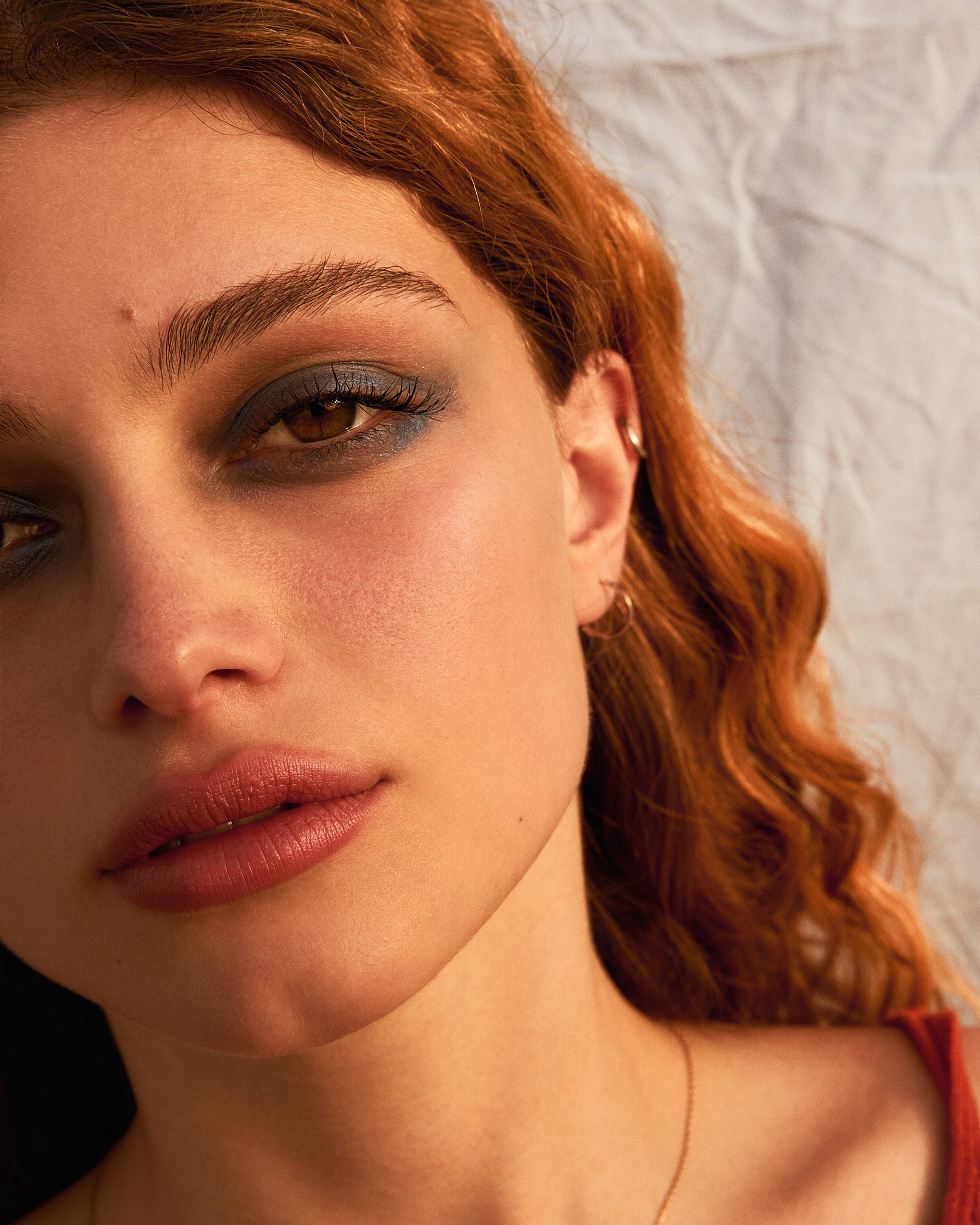 Close-up of a woman's face with red wavy hair, wearing makeup with smoky eyeshadow, and silver earrings, lying against a crumpled light-colored fabric background.