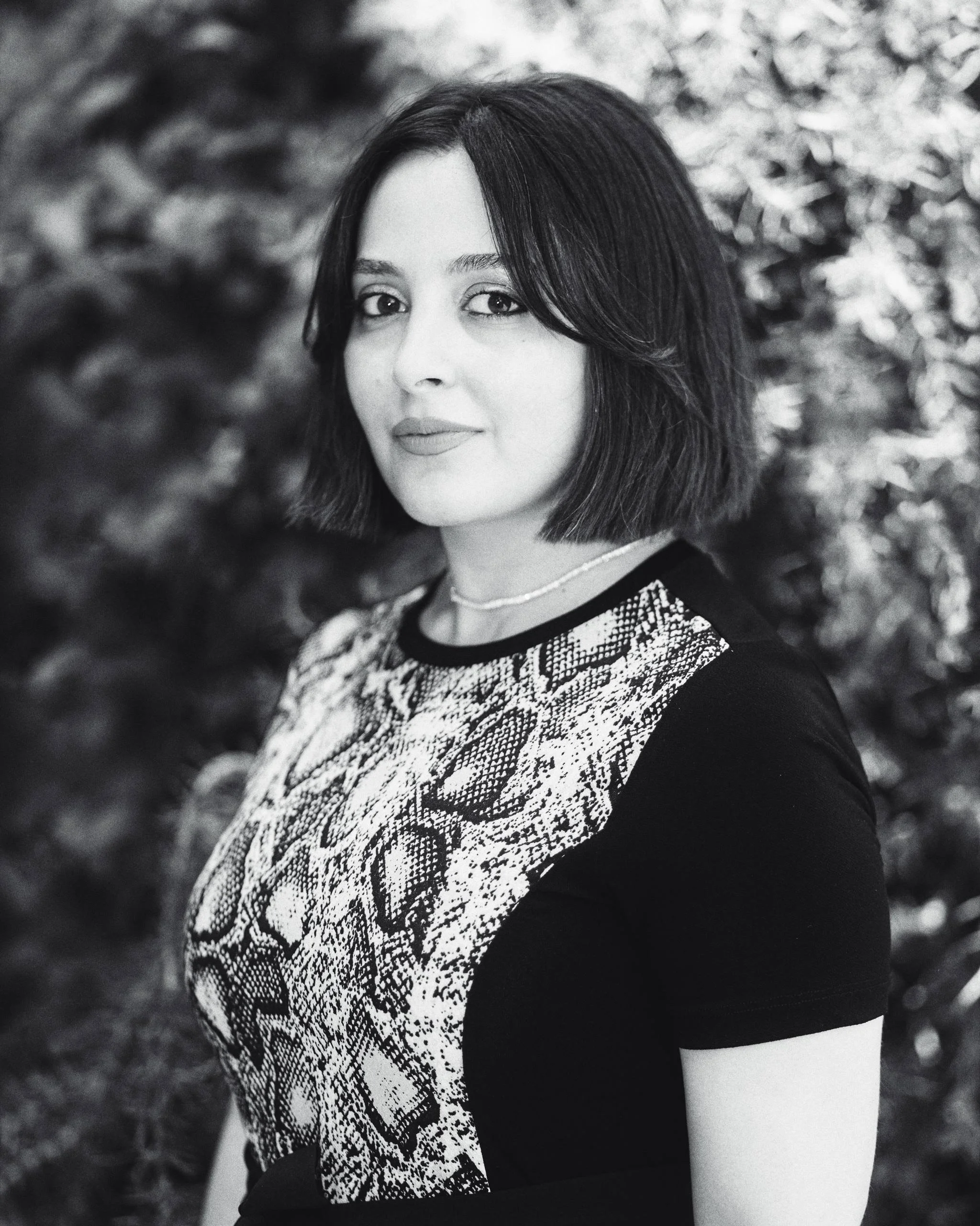 A woman with shoulder-length dark hair and light makeup, wearing a patterned top with snake skin design, a thin necklace, and a dark background of foliage.