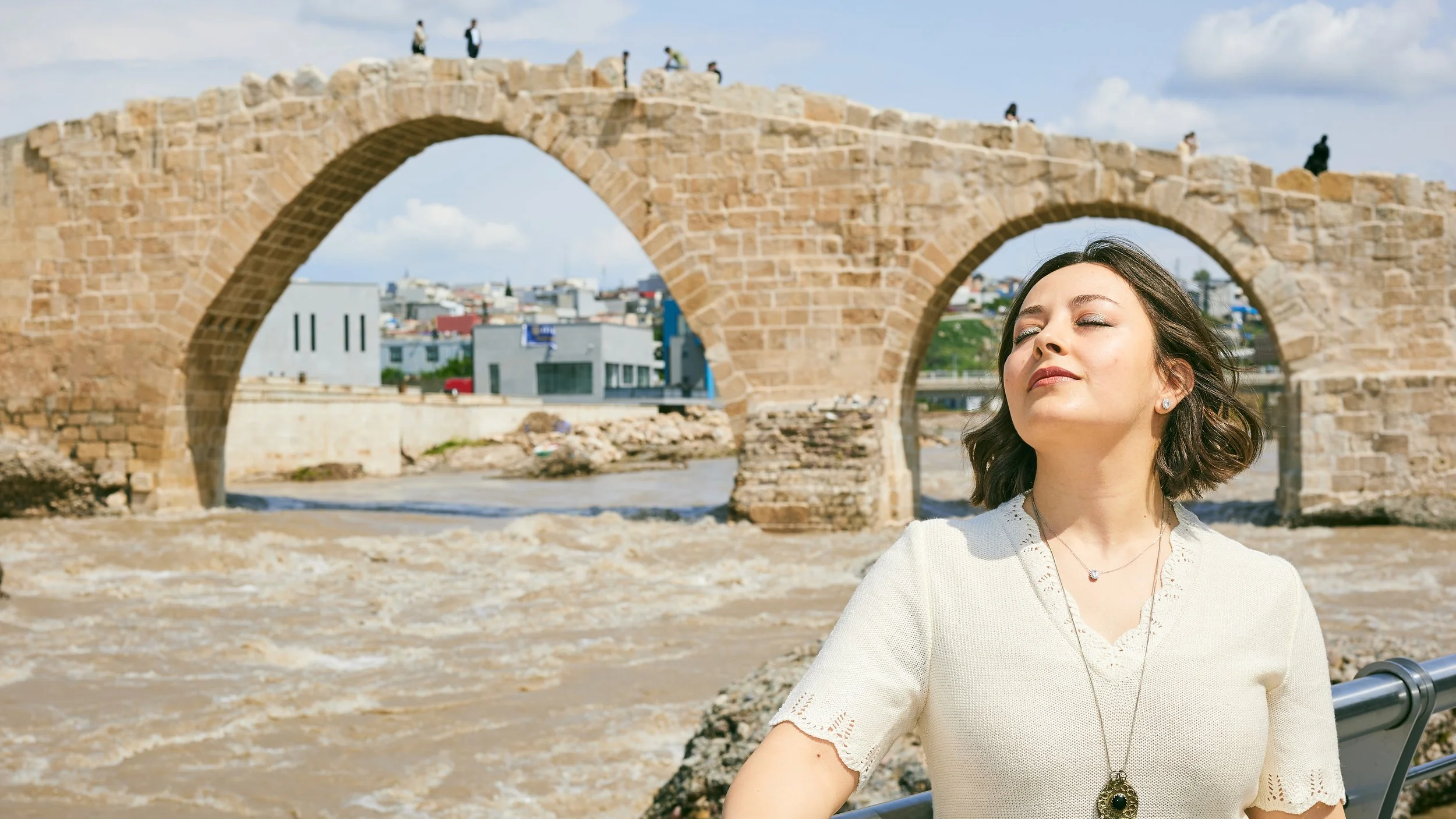 A woman with short dark hair and a white blouse stands with her eyes closed, enjoying the sunshine near an old stone bridge over a river with a city in the background.