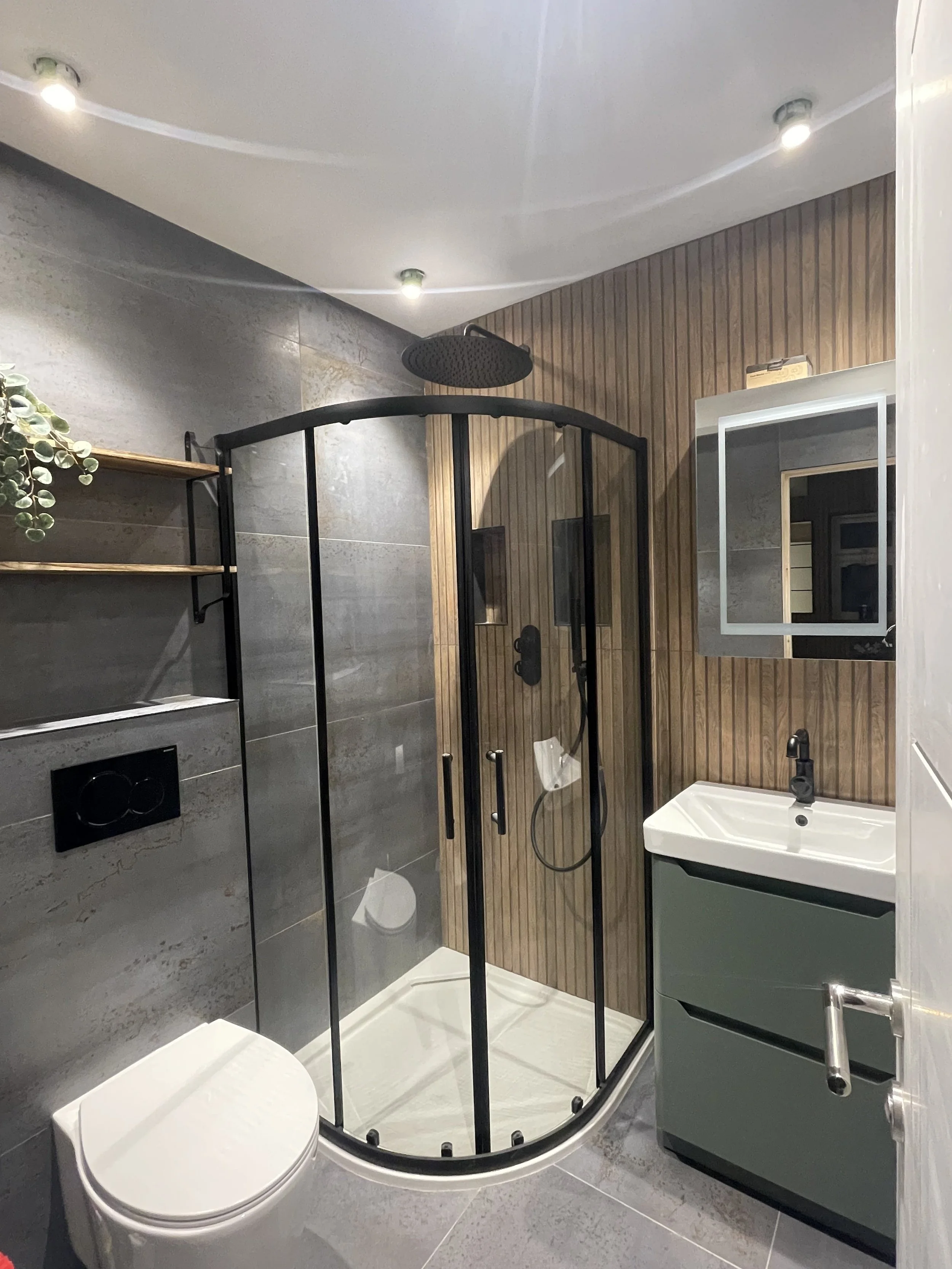 Modern bathroom with gray tiled walls, a glass shower enclosure with black framing, a white toilet, a small green vanity with a white sink, a mirror, and wood paneling on one wall.