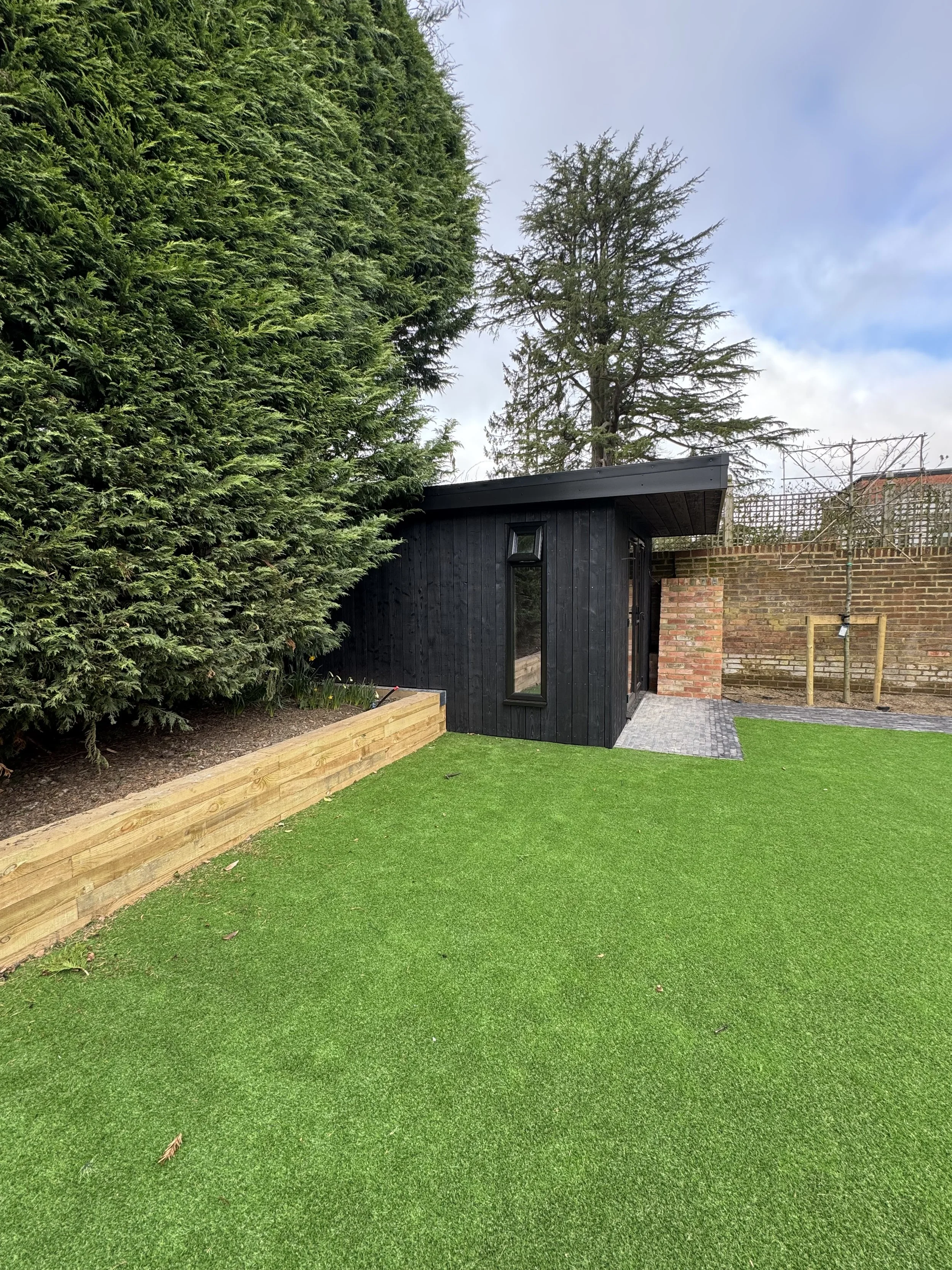 Small black wooden shed with tall narrow window, situated in a backyard with green artificial turf, a wooden border, shrubbery and trees, brick wall, and a paved pathway.
