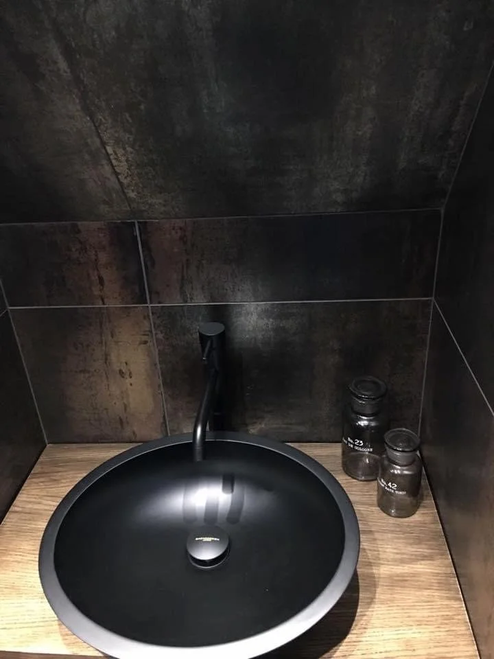 A black circular sink with a modern faucet mounted on a wooden countertop, with two small enameled jars labeled '42' and '23' beside it, set against a dark tiled wall.