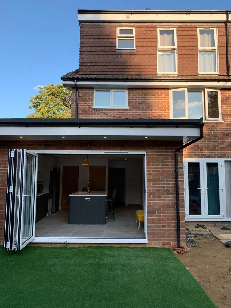 The back of a brick house with open sliding glass doors revealing a modern kitchen and living area, with a small patch of grass in the foreground and a clear blue sky above.