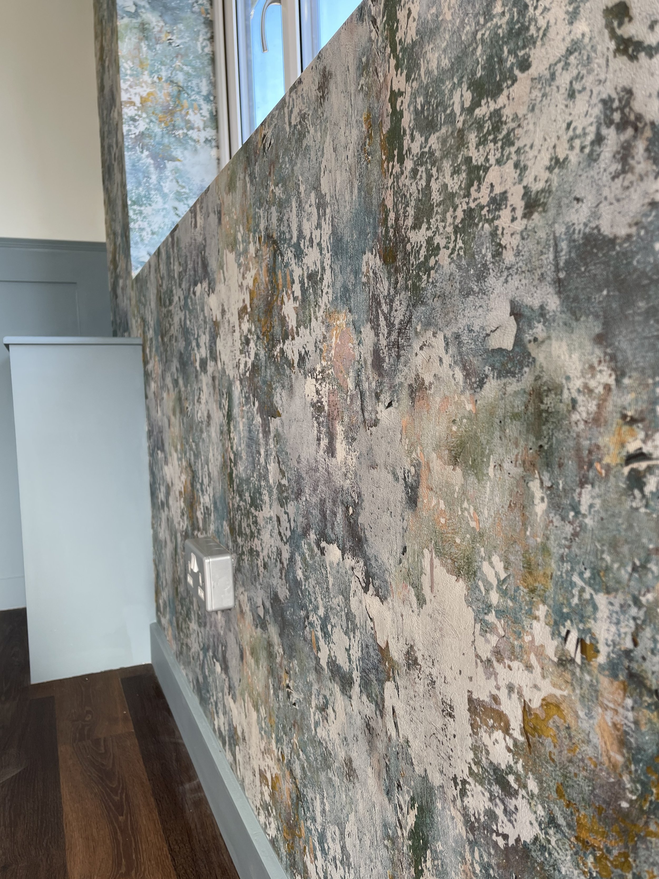 Close-up of a textured, multicolored abstract wall surface with a visible electrical outlet, indoors with a window in the background.