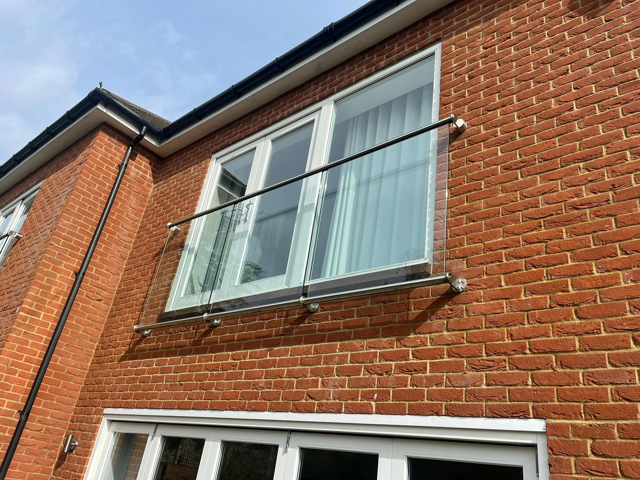 A balcony with glass railing on a brick house, above large glass sliding doors, under a black tiled roof with a black rain gutter.