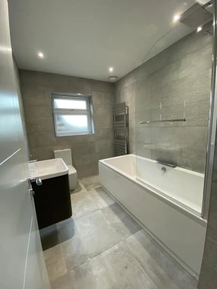 Modern bathroom with gray tiled walls and floors, a white bathtub with a glass shower screen, a white toilet, a grey sink vanity, a small window, and a heated towel rail.