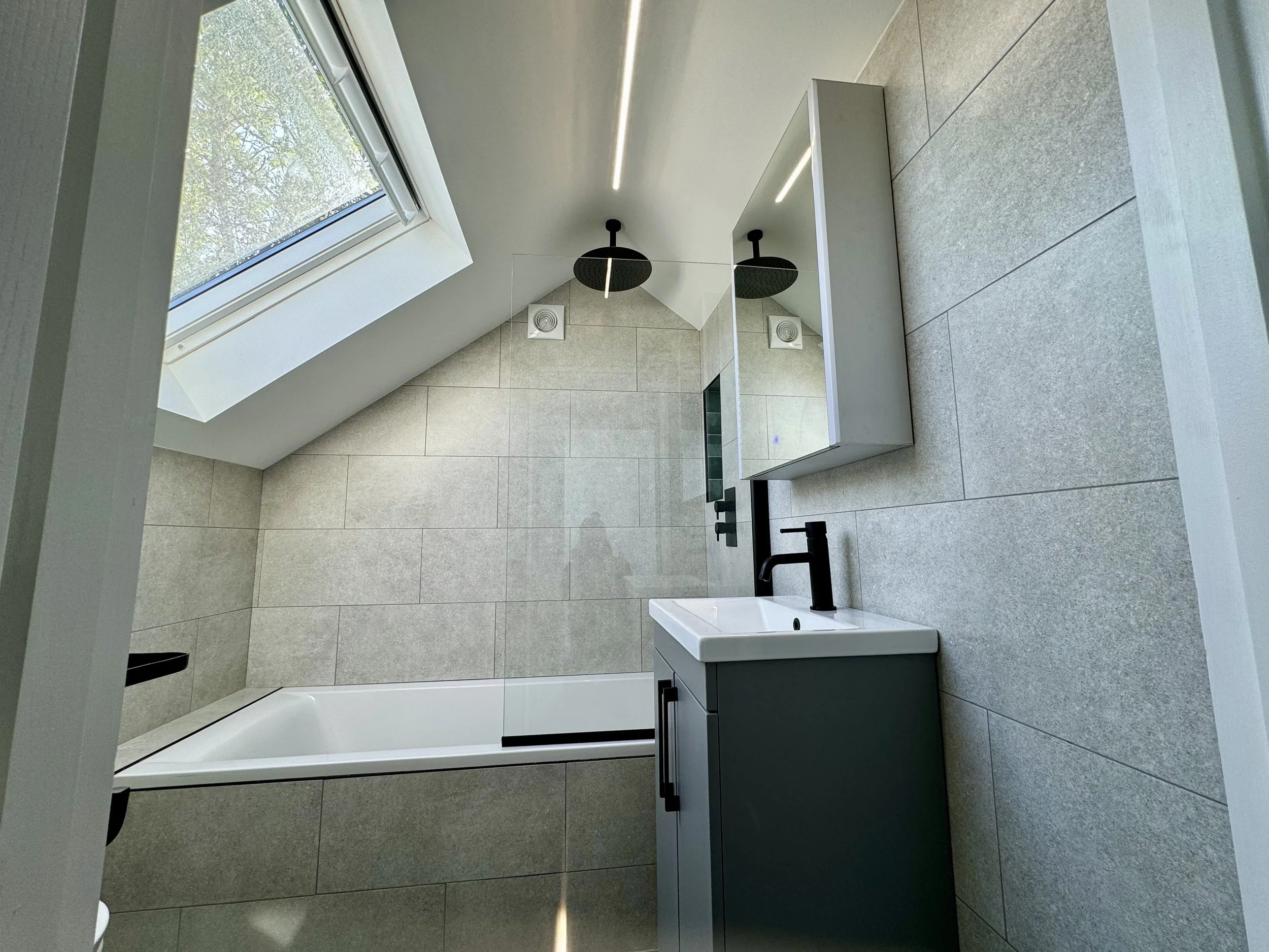 Modern bathroom featuring a skylight window, a black showerhead, a small sink with a black faucet, a mirrored cabinet, and beige tiled walls and floor.