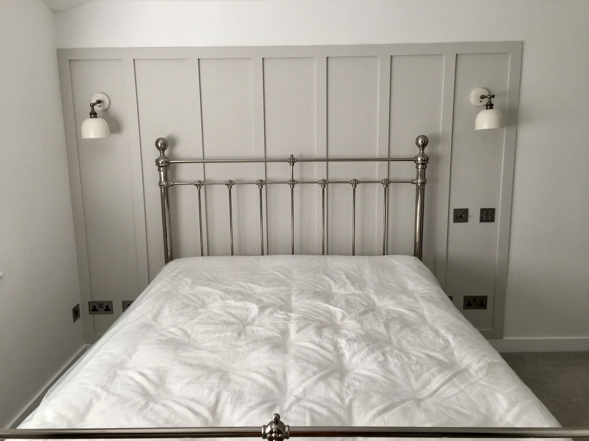 A bedroom with a metal bed frame and a white mattress, white walls, wall-mounted lamps, and electrical outlets.