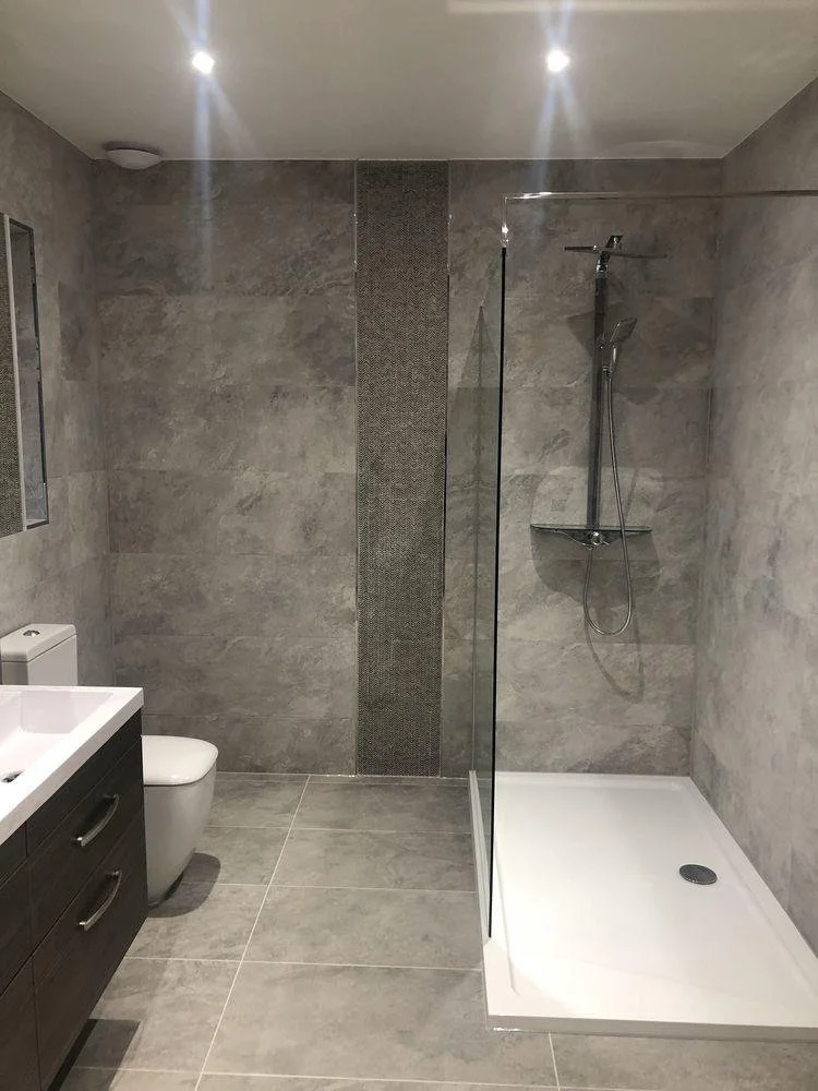 Modern bathroom with gray tiled walls and floor, a white toilet, a dark wood vanity with a white sink, a mirror, and a walk-in shower with a glass partition, a silver shower head, and a white base.