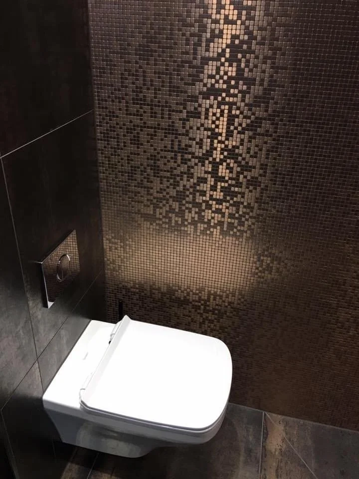 A modern bathroom with a white wall-mounted toilet and a bronze mosaic tiled wall behind it.
