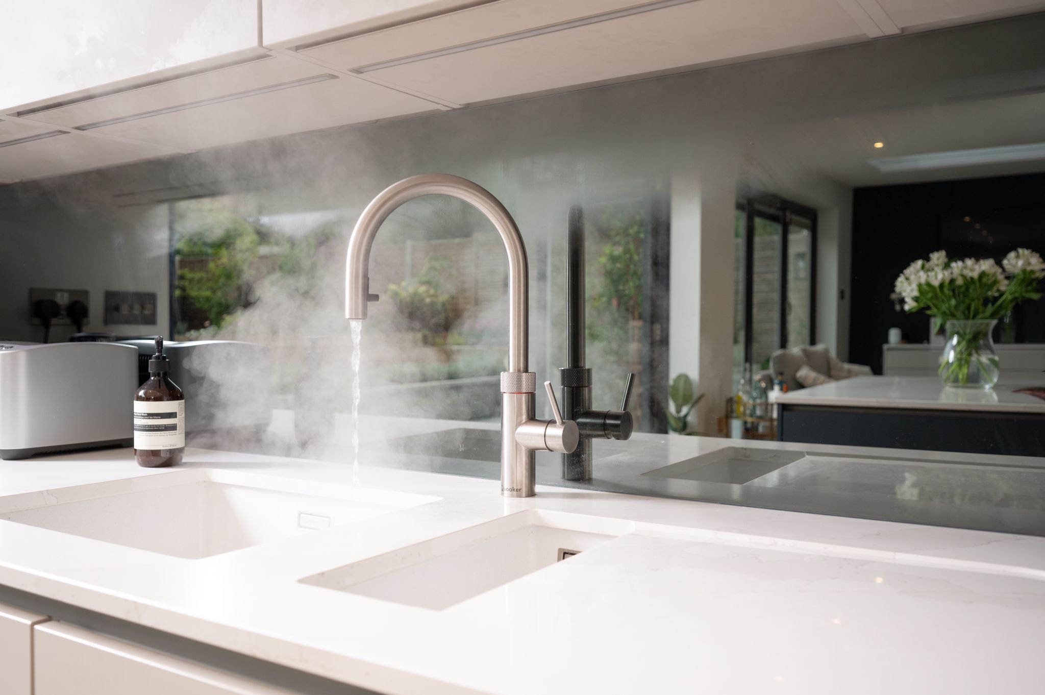 Modern kitchen sink with water running from a stainless steel faucet, steam rising from the hot water, and a soap dispenser on the white countertop, with a view into a living room with flowers in a vase.